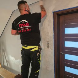 Wykończenia Wnętrz Marcin Wójcik - Pracownik na podeście aplikuje dekoracyjną szpachlę na ścianie wewnątrz pomieszczenia, widoczne zabezpieczone taśmą drzwi i gniazdka elektryczne.