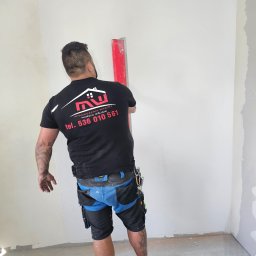 Wykończenia Wnętrz Marcin Wójcik - Pracownik w czarnej koszulce z logo firmy wykończeniowej, wyrównuje ścianę czerwoną łatą. Widoczne ślady gładzi na ścianie i podłodze. W tle narożnik pokoju.