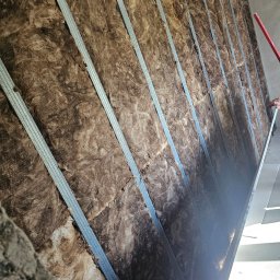 Wykończenia Wnętrz Marcin Wójcik - Izolacja ścian wełną mineralną, widoczne metalowe profile konstrukcyjne. Prace wykończeniowe w trakcie montażu. Ujęcie z dołu, ukazujące strukturę materiałów.