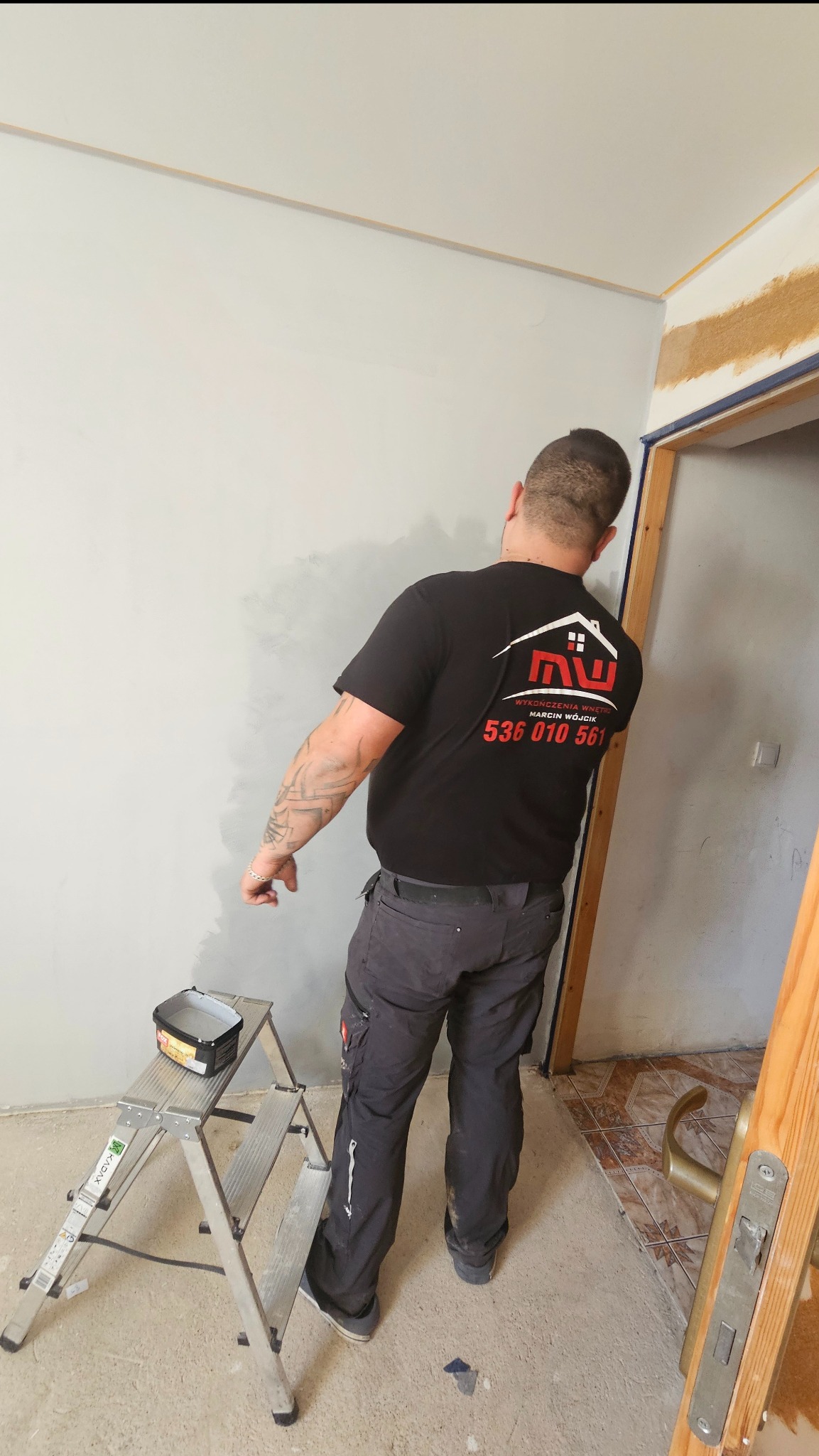 Mężczyzna w czarnej koszulce z logo firmy maluje szarą farbą ścianę wewnątrz pomieszczenia. Widoczna drabina i puszka z farbą. Częściowo wykończone wnętrze.