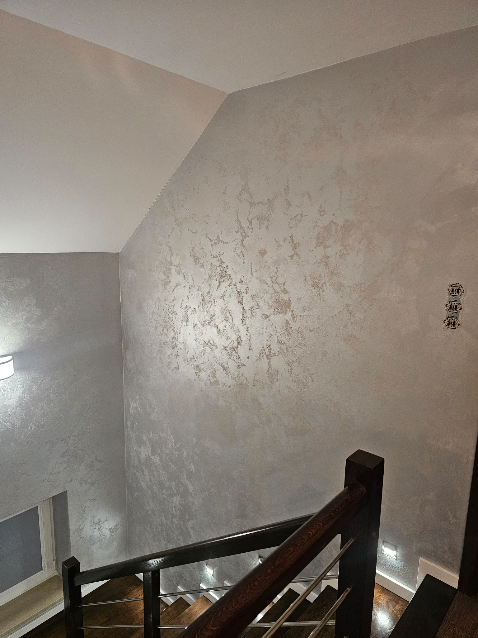 Ściana w korytarzu wykończona dekoracyjnym tynkiem w odcieniach szarości i beżu, widok z góry na schody z drewnianą balustradą i oświetleniem punktowym.