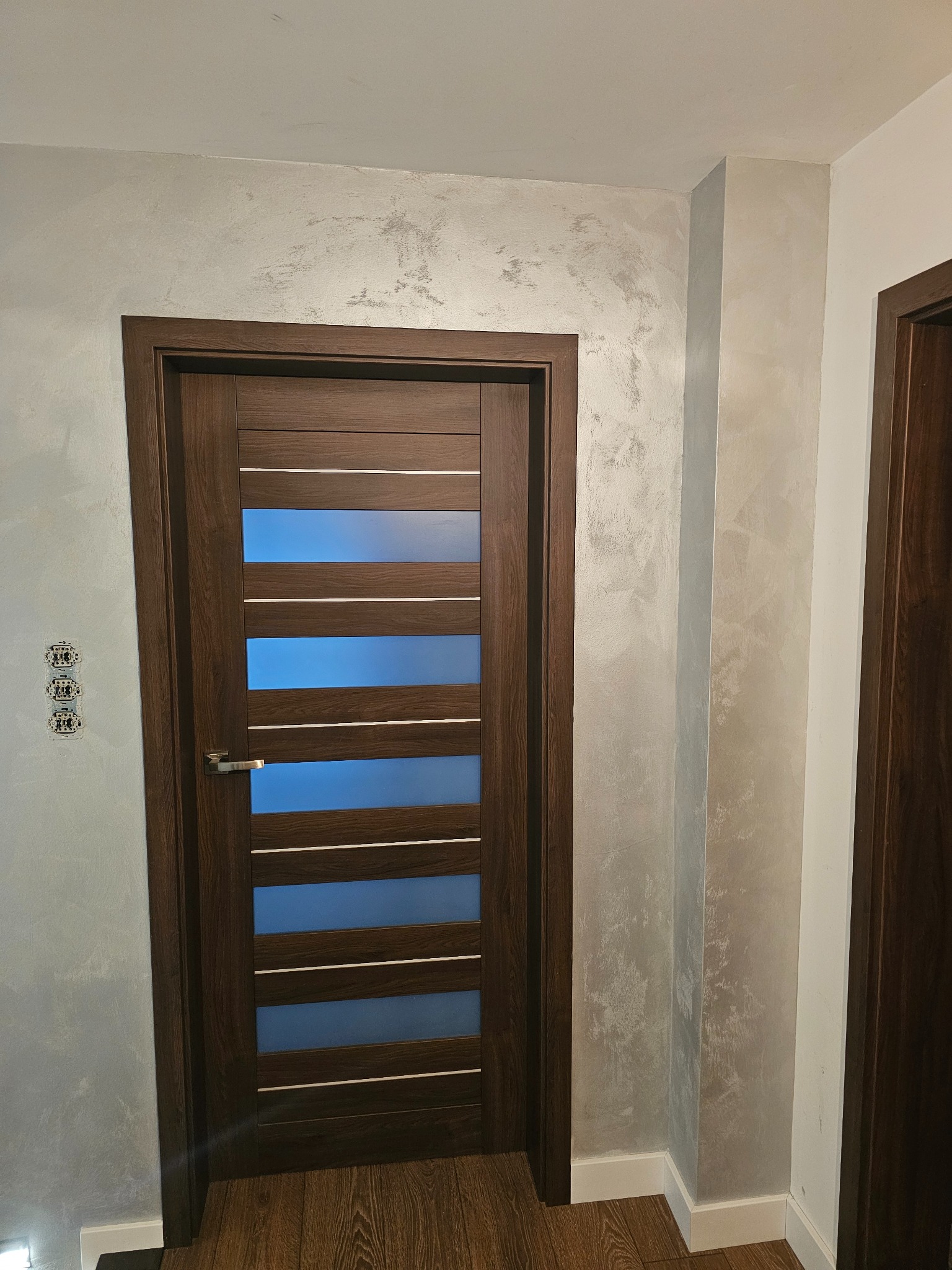 Nowoczesne drzwi wewnętrzne z przeszkleniami w brązowej ramie, zamontowane w ścianie o dekoracyjnym wykończeniu. Widoczne gniazdka elektryczne i fragment podłogi.