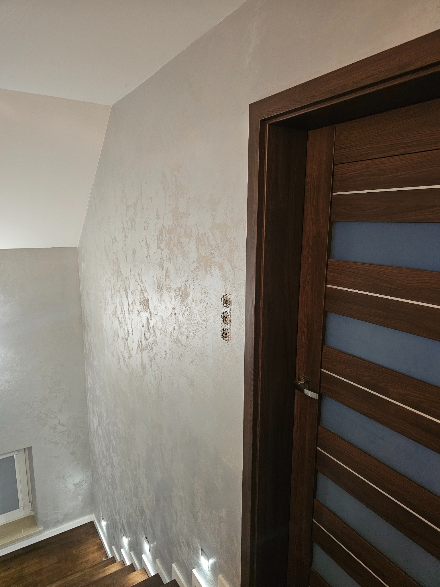Wykończona ściana z dekoracyjnym tynkiem w odcieniach szarości, widoczne schody z oświetleniem podstopnic, drewniane drzwi z przeszkleniami i włączniki światła.