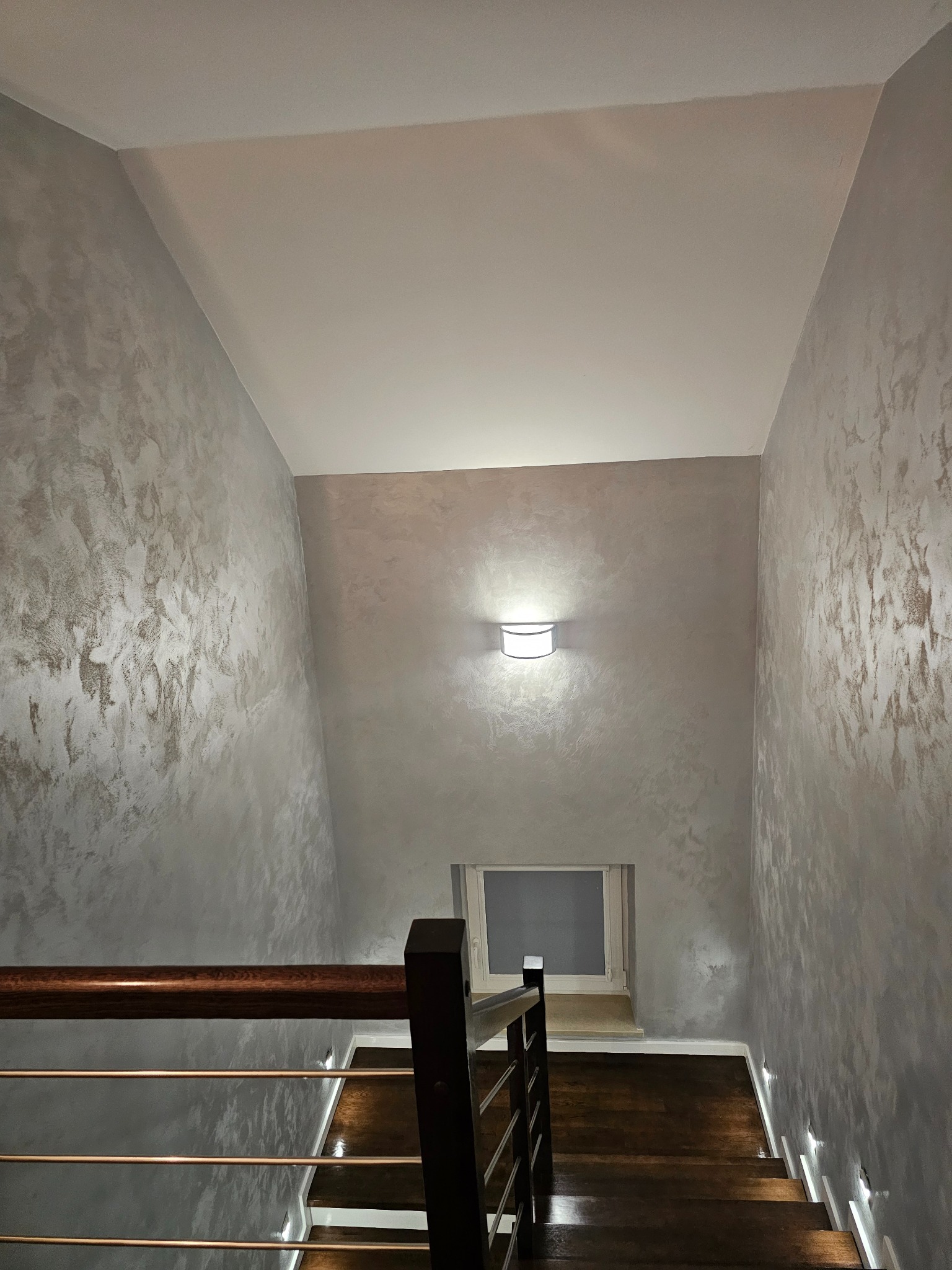 Klatka schodowa z drewnianymi schodami i metalową balustradą. Ściany wykończone dekoracyjnym tynkiem w odcieniach szarości. Oświetlenie schodów punktowe, dodatkowo lampa na ścianie.