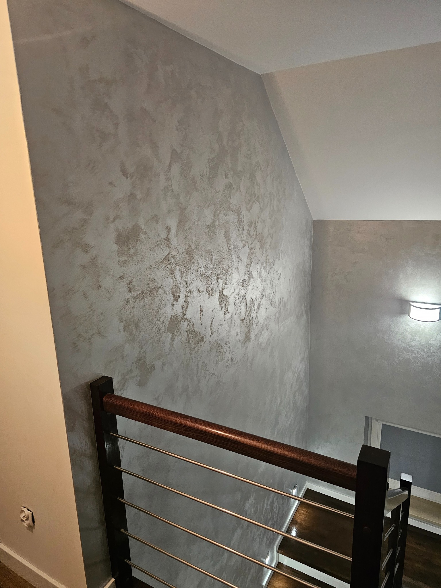Dekoracyjna ściana w odcieniach szarości z efektem stiuku w klatce schodowej, widok z góry na drewniane schody i metalową balustradę.