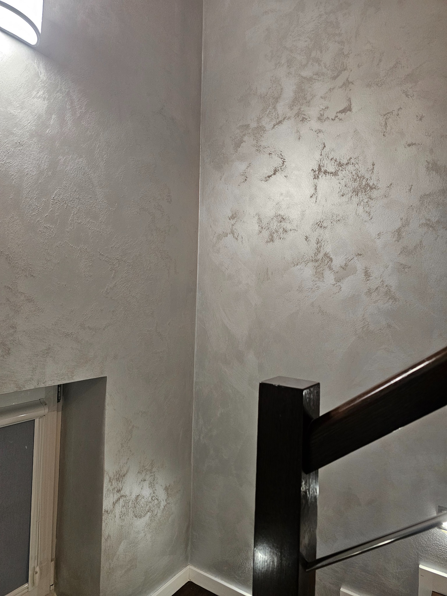 Ściana wewnętrzna ze szpachlowaniem dekoracyjnym w odcieniach szarości i beżu, widoczna fragment schodów z drewnianą balustradą i okno z roletą.