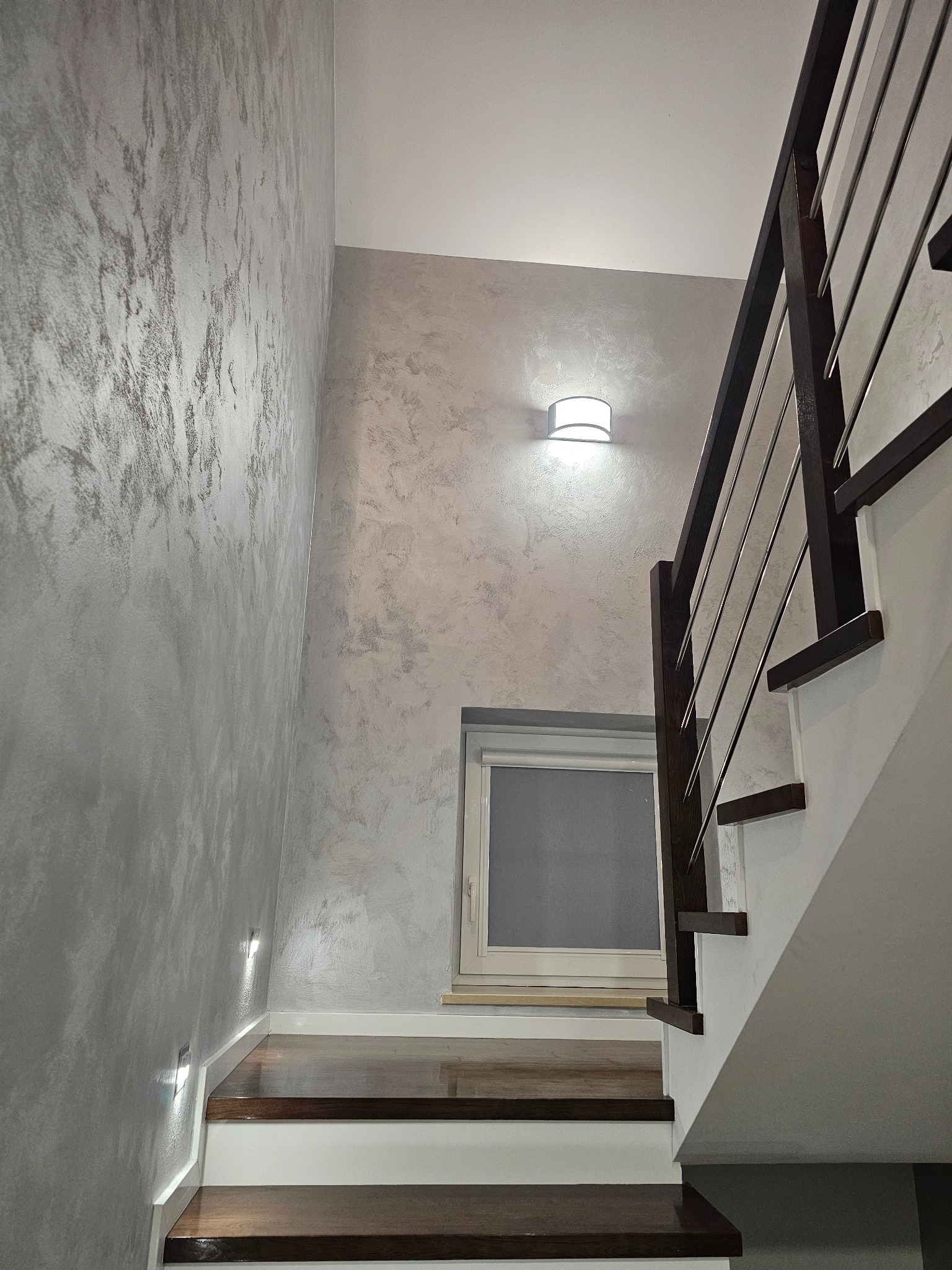 Klatka schodowa z dekoracyjną szarą ścianą, oświetleniem punktowym i drewnianymi schodami. Widoczne okno i fragment balustrady. Nowoczesny design.