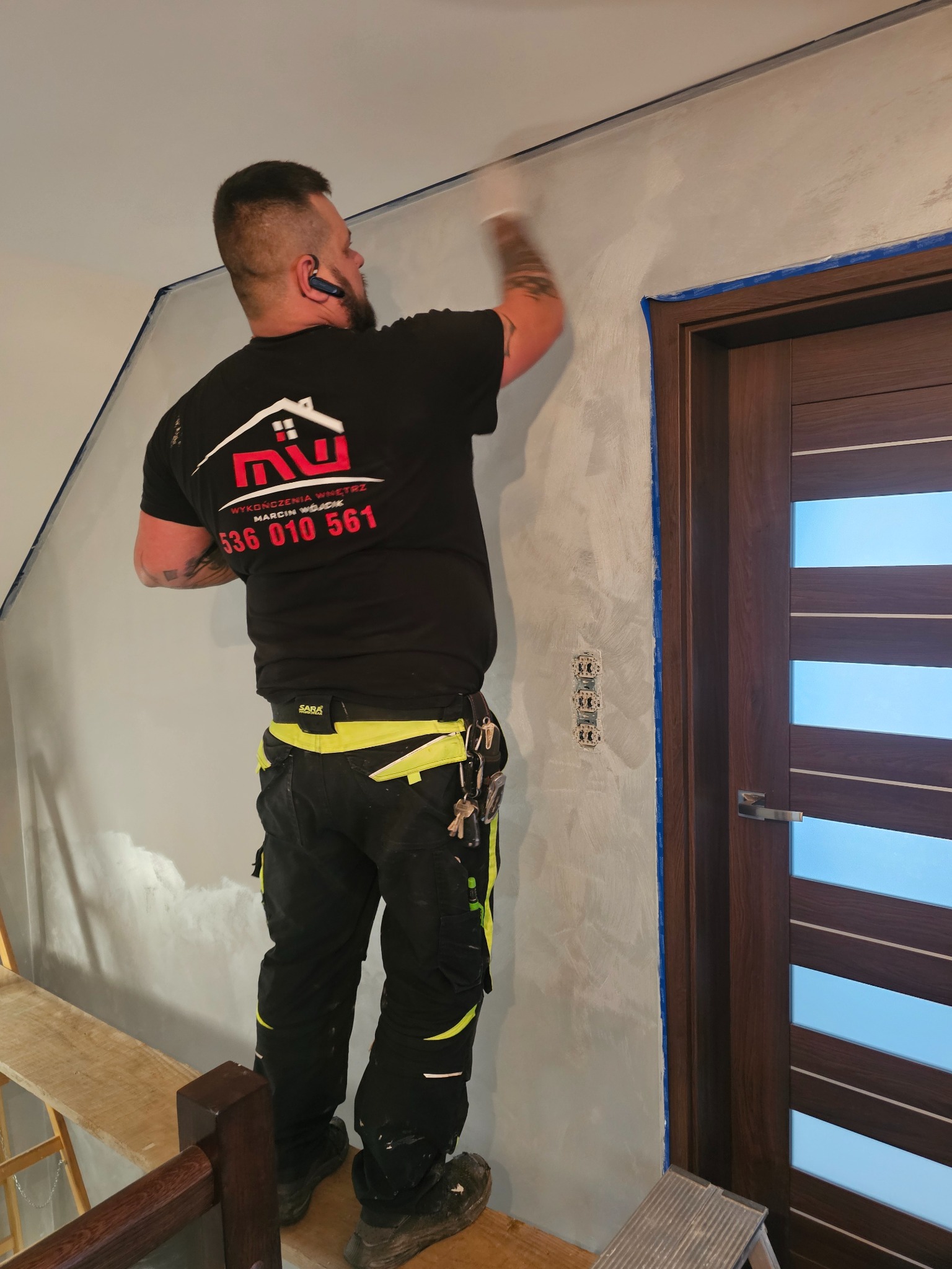 Pracownik na podeście aplikuje dekoracyjną szpachlę na ścianie wewnątrz pomieszczenia, widoczne zabezpieczone taśmą drzwi i gniazdka elektryczne.