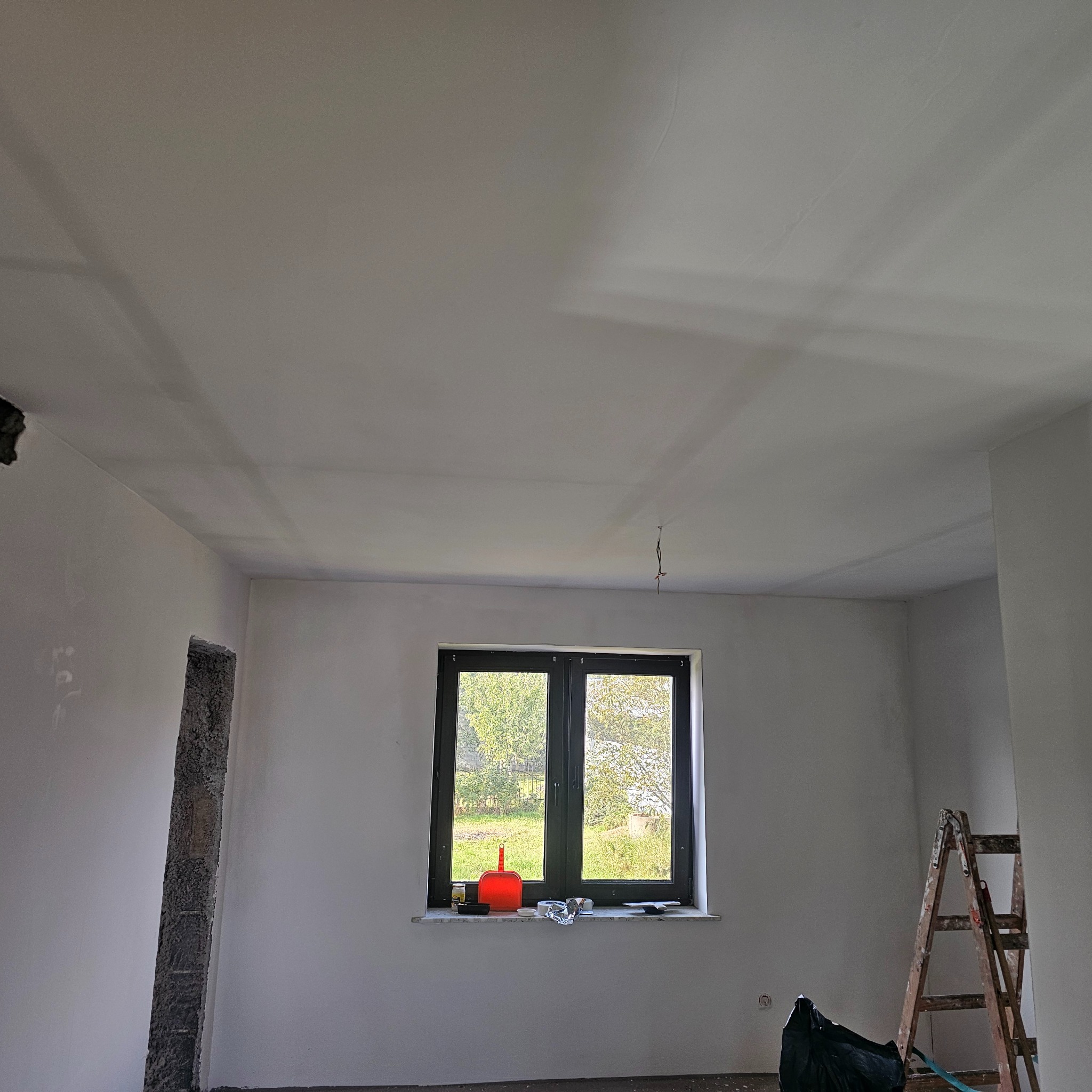 Wnętrze po gładziowaniu ścian i sufitu, widoczne okno z parapetem, drabina malarska i otwór w ścianie. Ściany pomalowane na biało, przygotowane do dalszych prac.