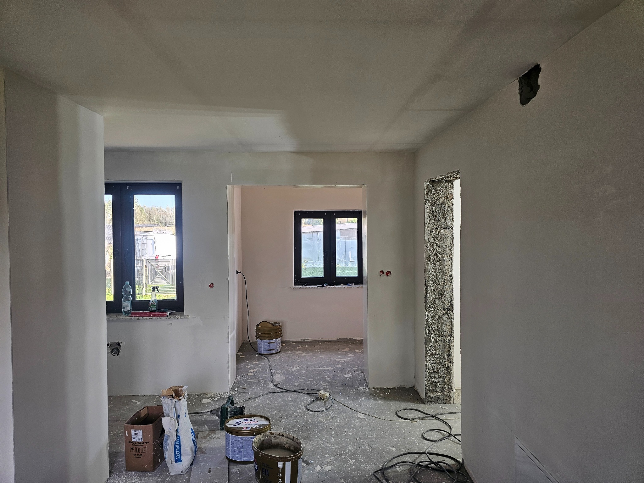 Wnętrze po nałożeniu gładzi, widoczne narzędzia i materiały budowlane. Otwór w suficie. Dwa okna z czarną ramą w tle. Stan surowy, przygotowanie do dalszych prac wykończeniowych.