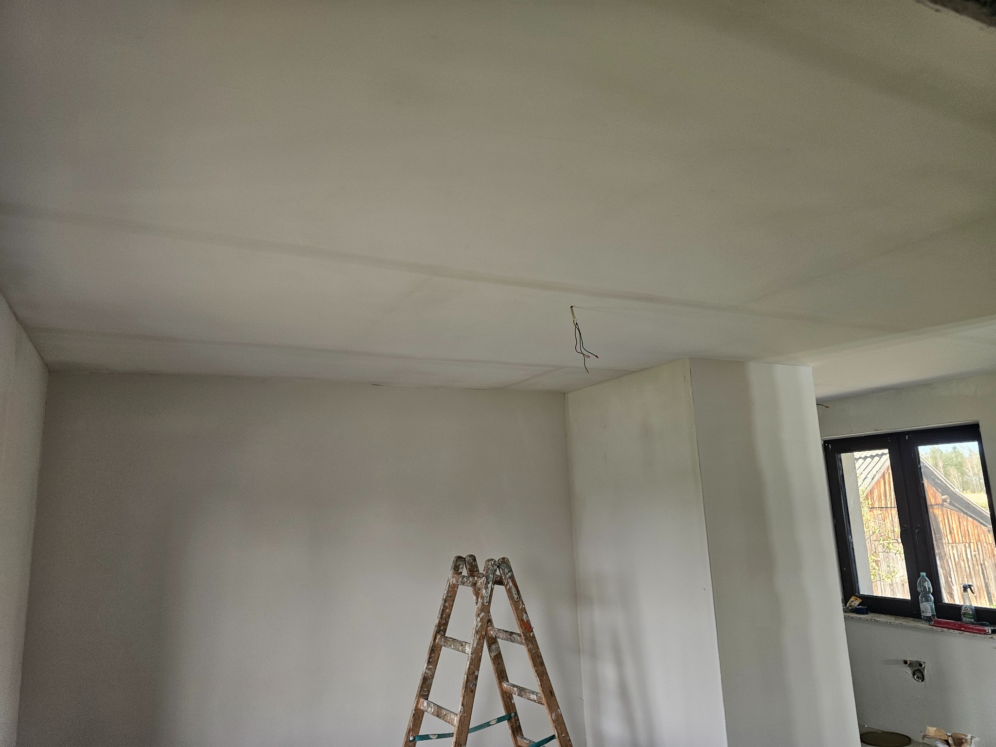 Wnętrze po gładziowaniu: białe ściany i sufit, drewniana drabina malarska, okno z widokiem na zewnątrz. Widoczne ślady po pracach wykończeniowych.