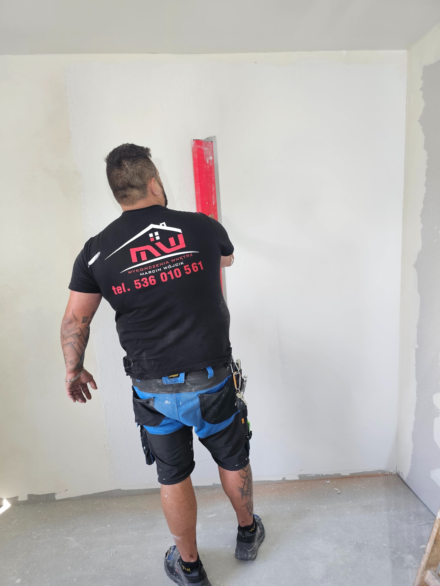 Pracownik w czarnej koszulce z logo firmy wykończeniowej, wyrównuje ścianę czerwoną łatą. Widoczne ślady gładzi na ścianie i podłodze. W tle narożnik pokoju.