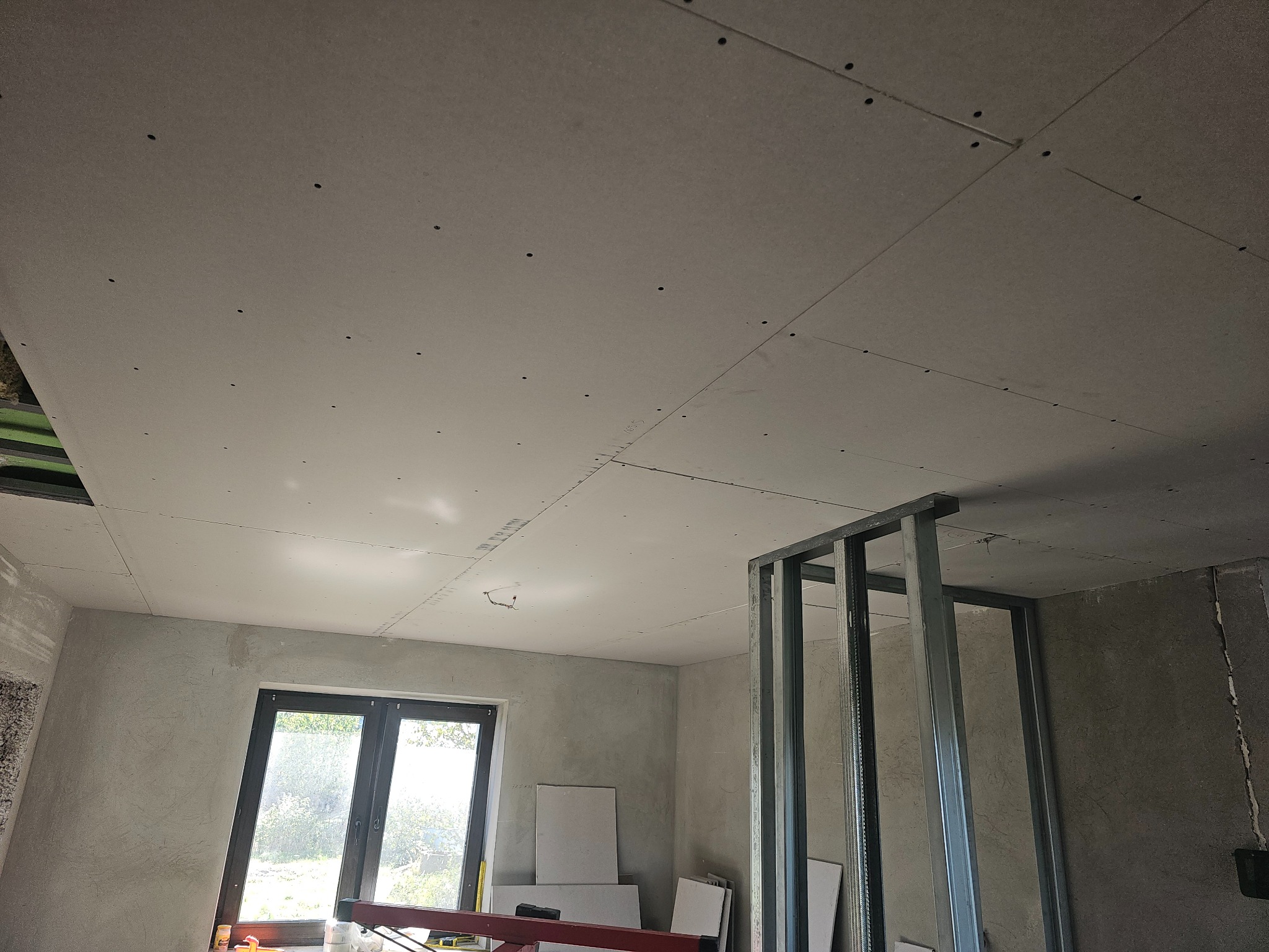 Montaż płyt gipsowo-kartonowych na suficie w surowym wnętrzu. Widoczne metalowe profile konstrukcyjne i okno z widokiem na zieleń. Etap prac wykończeniowych.