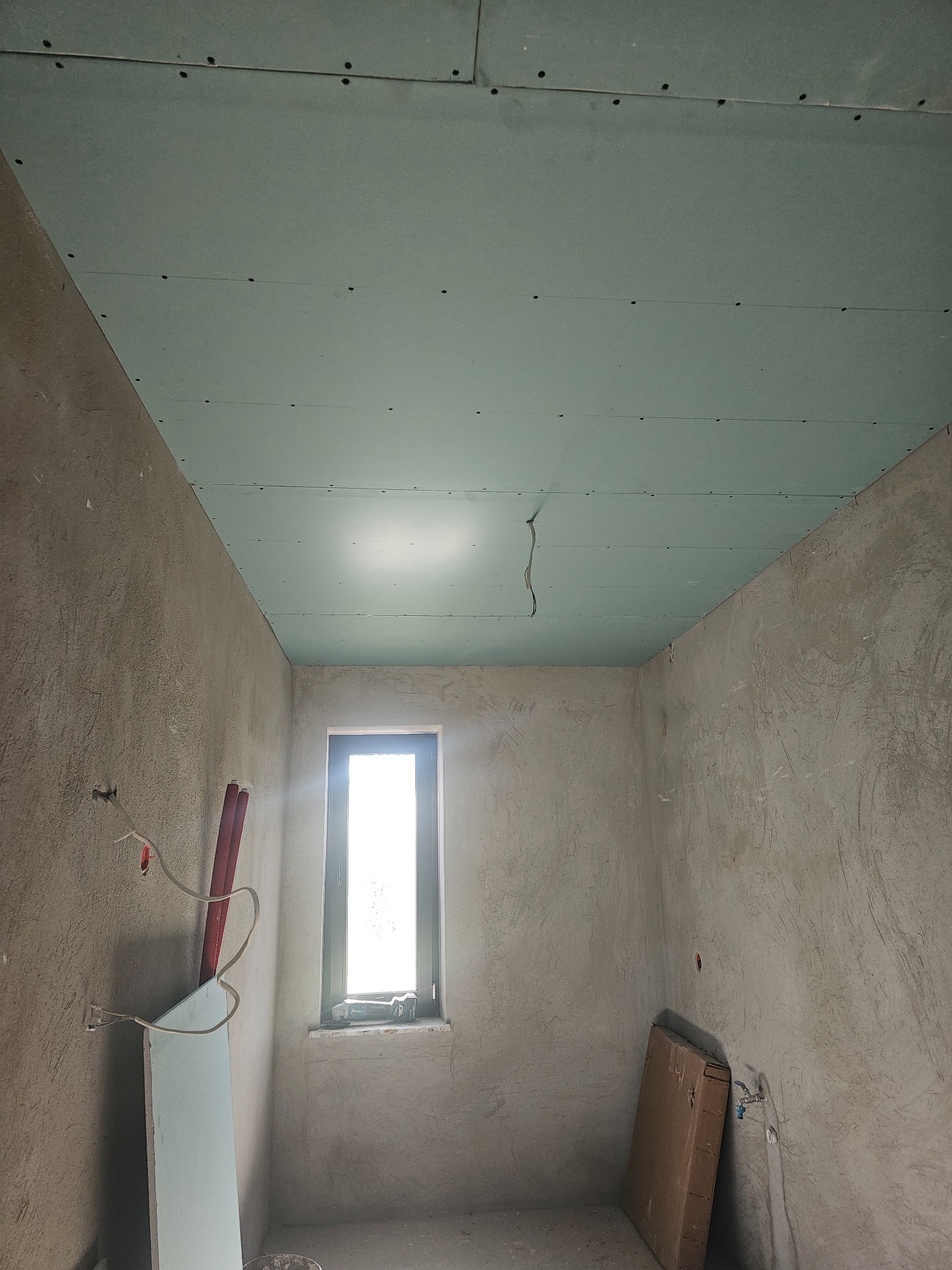 Montaż sufitu podwieszanego z płyty karton-gips w surowym pomieszczeniu z oknem. Widoczne przewody i rury instalacyjne na ścianach.