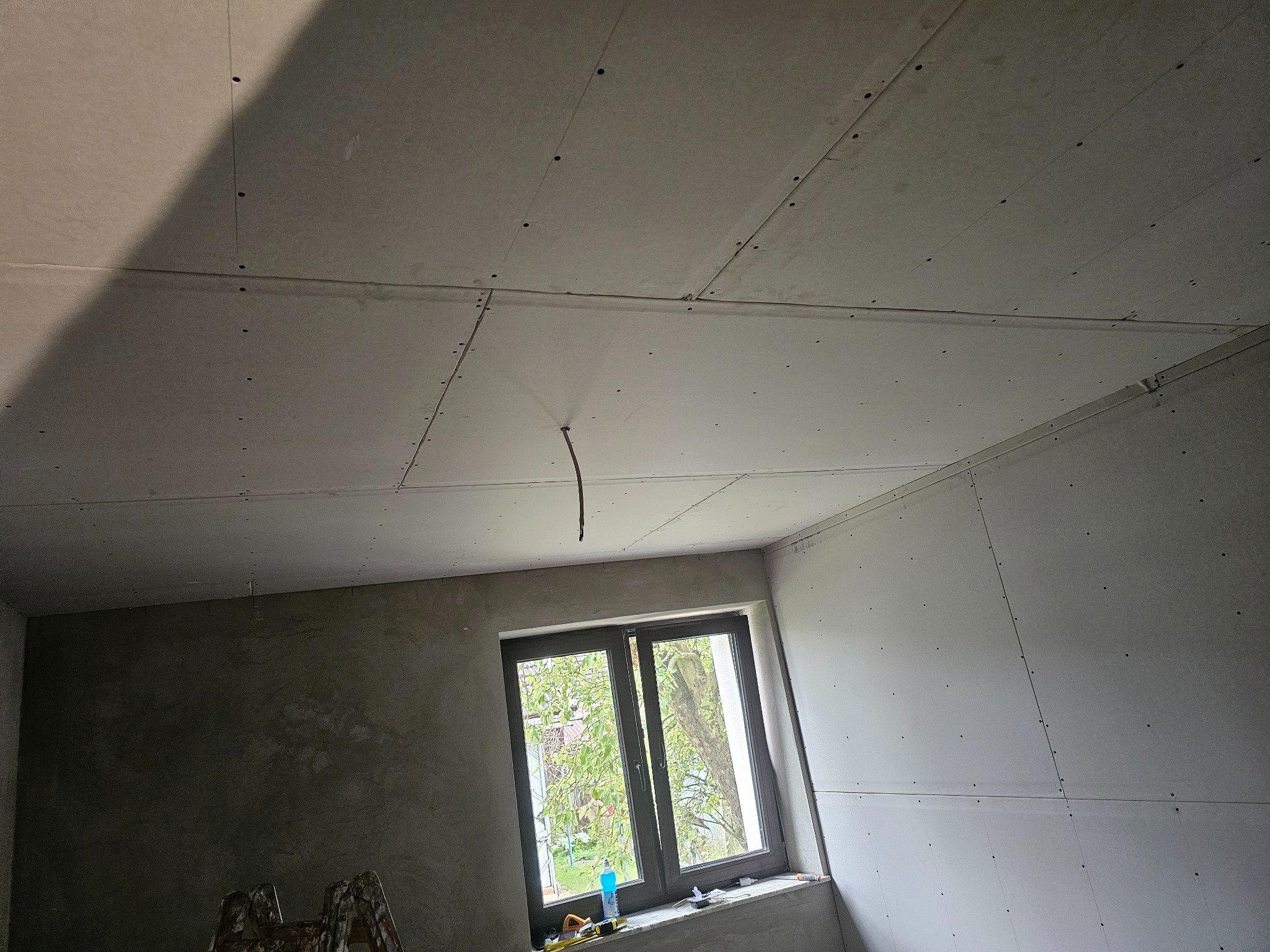 Pokój w trakcie montażu płyt kartonowo-gipsowych na suficie i ścianach. Widoczne łączenia płyt, wystający kabel i drabina malarska. Okno z widokiem na zieleń.