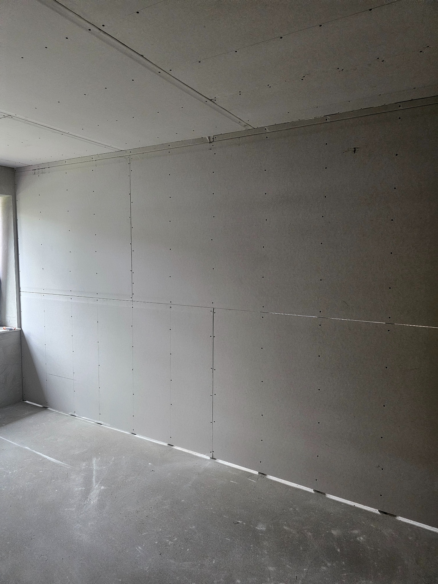 Pokój w trakcie montażu płyt kartonowo-gipsowych na ścianach i suficie. Widoczne łączenia płyt i szpachlowane wkręty. Surowy beton na podłodze, okno z lewej strony.