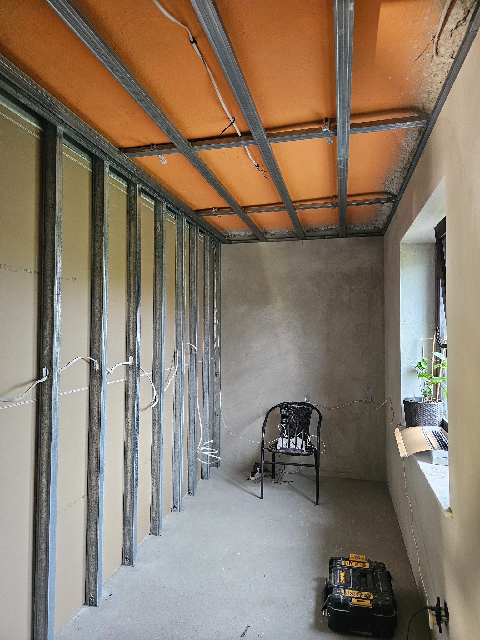 Montaż płyt karton-gips na ścianie i suficie z widoczną konstrukcją metalową. W tle surowa ściana i okno z rośliną. Na podłodze skrzynka narzędziowa.