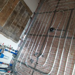 NGP Sp. z o.o. - Instalacja ogrzewania podłogowego z białych rur na folii z czerwoną kratką, widoczne elementy hydrauliczne i fragment drewnianej podłogi w tle.