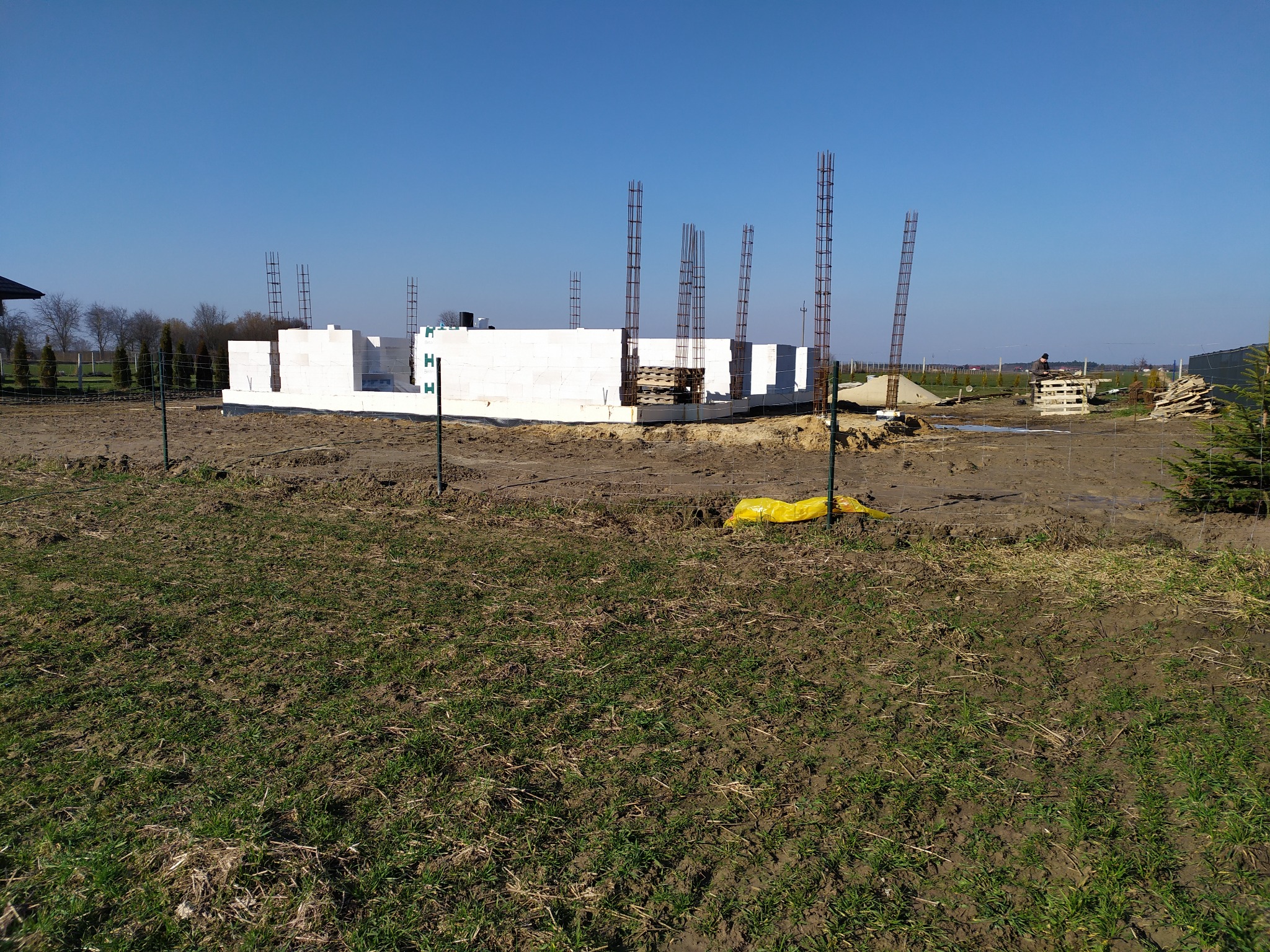 Wznoszenie ścian z białych bloczków na fundamencie z wystającymi prętami zbrojeniowymi. Widok na plac budowy w słoneczny dzień.