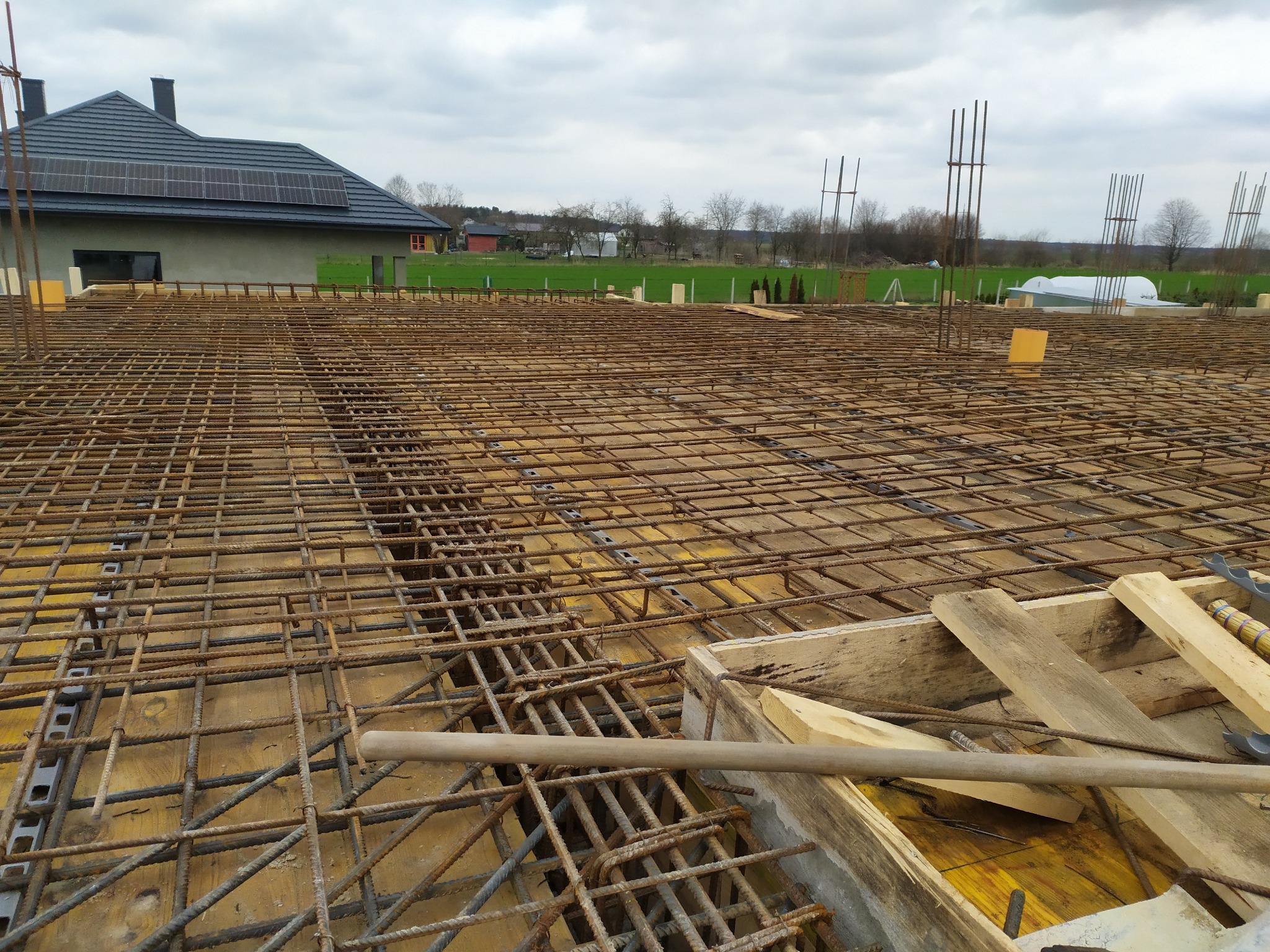 Zbrojenie fundamentów domu jednorodzinnego z widoczną konstrukcją szalunkową i wystającymi prętami. W tle dom z panelami słonecznymi i zielony krajobraz.