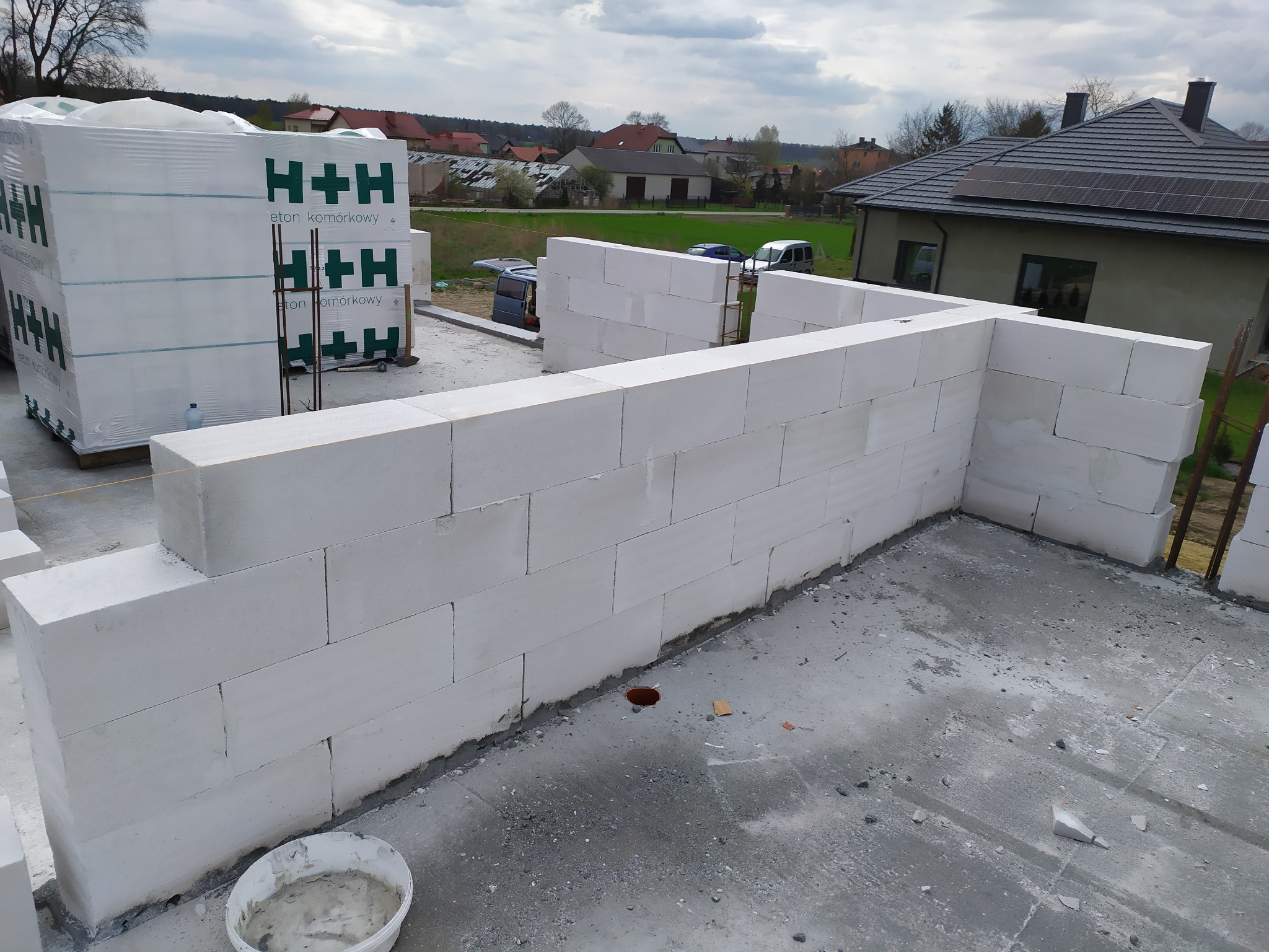 Wznoszenie ścian z betonu komórkowego H+H na fundamencie domu jednorodzinnego. Widoczne palety z bloczkami i wiadro z zaprawą. W tle zabudowa jednorodzinna.