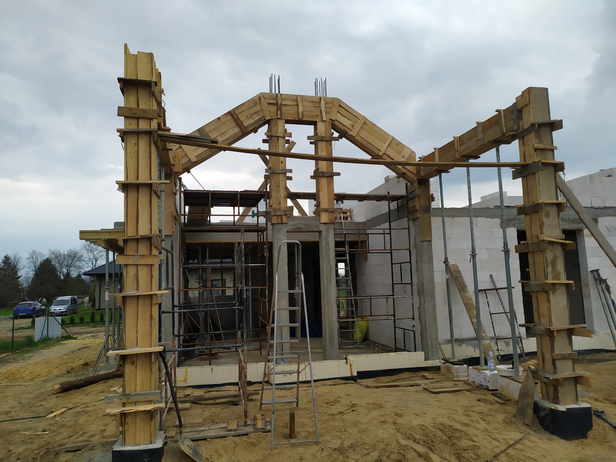 Budowa domu w trakcie: szalunki drewniane kolumn i nadproży, ściany z betonu komórkowego, drabina, widok na budowę w pochmurny dzień.