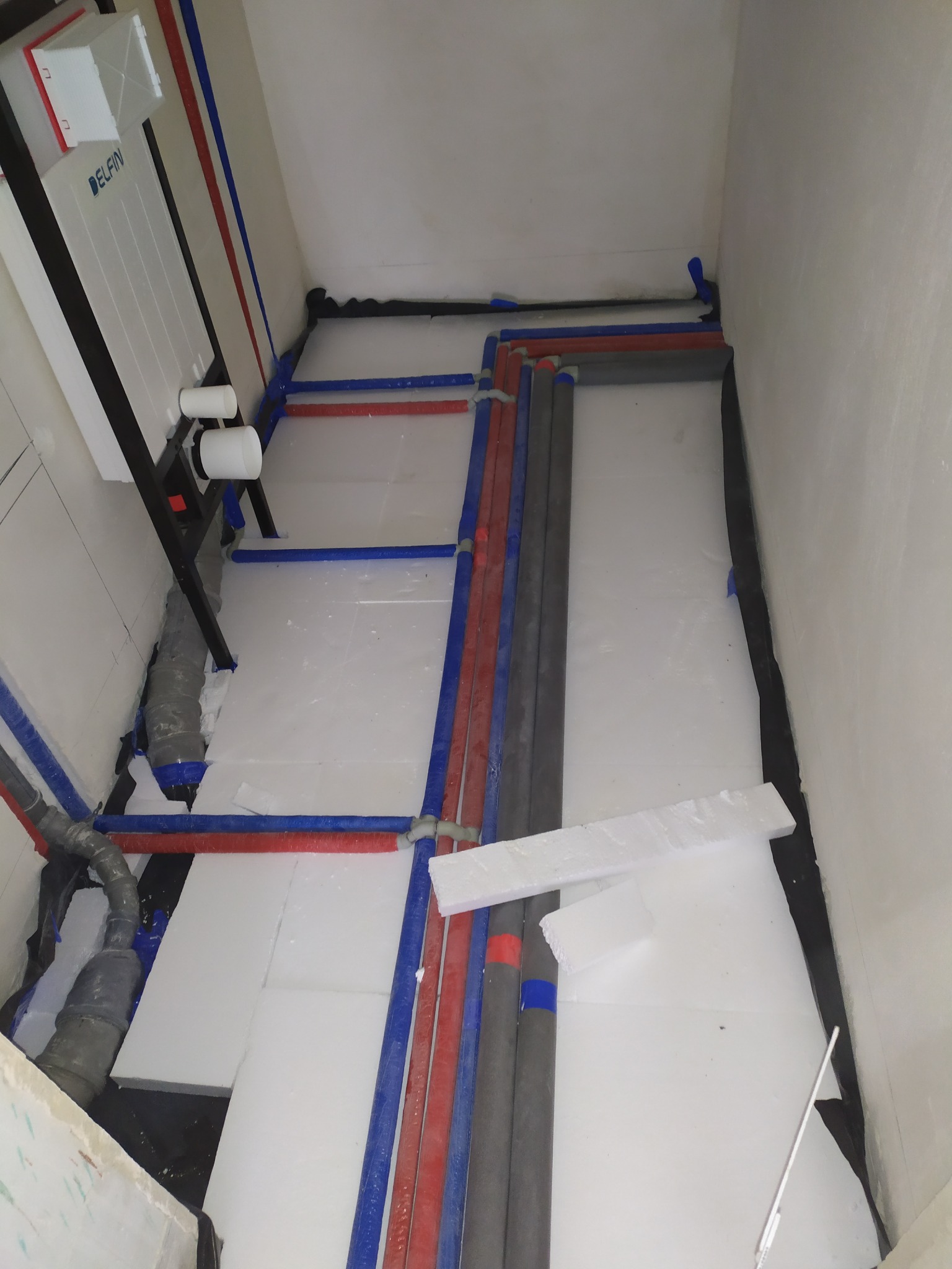 Instalacja podtynkowa WC z rurami wodnymi (czerwone i niebieskie) oraz kanalizacyjnymi, ułożona na białym styropianie. Widok z góry, surowy stan.