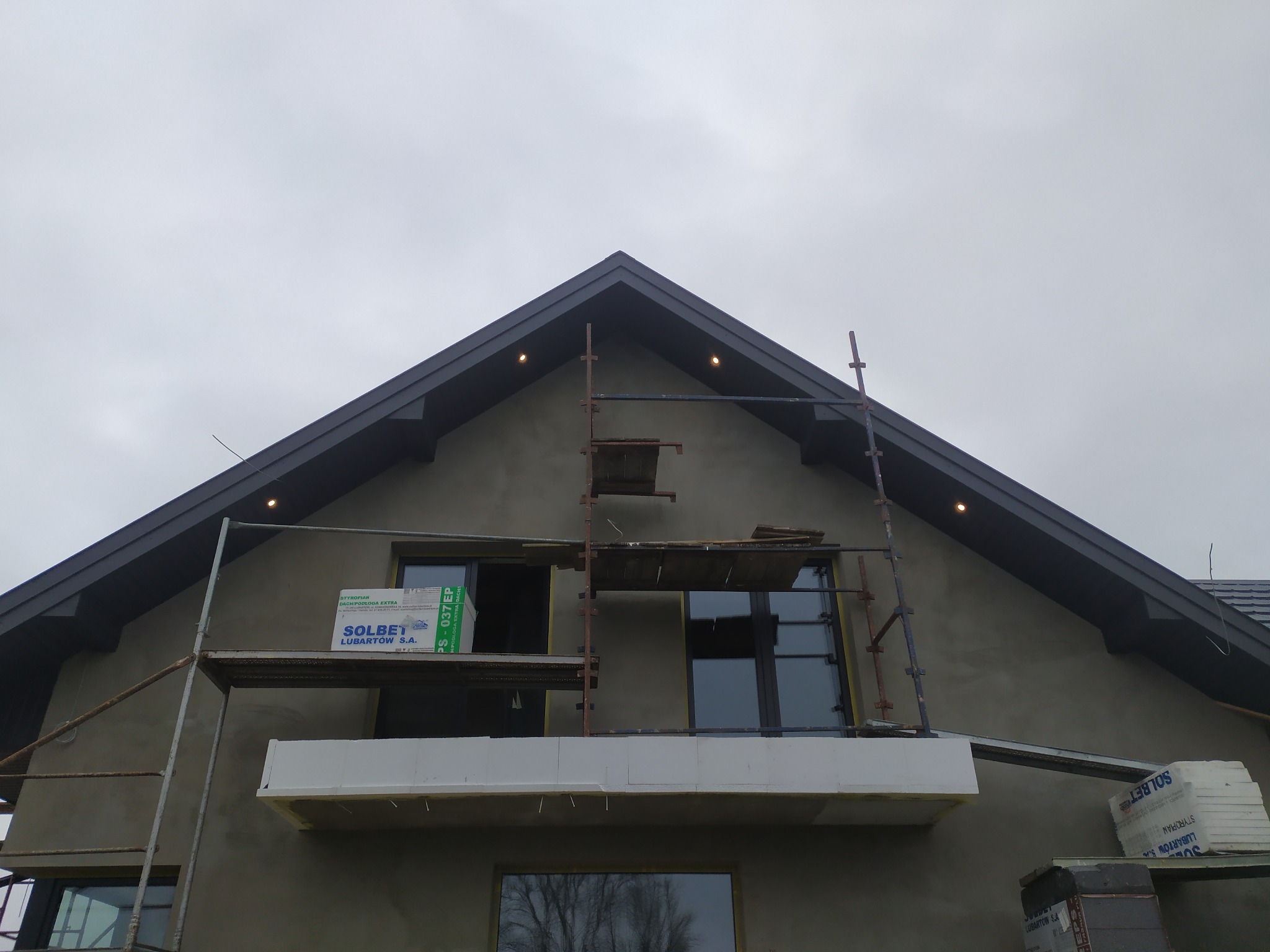 Dom w trakcie ocieplania, widoczne rusztowania, styropian Solbet i świeżo nałożony tynk. Ujęcie z dołu, pochmurne niebo w tle.