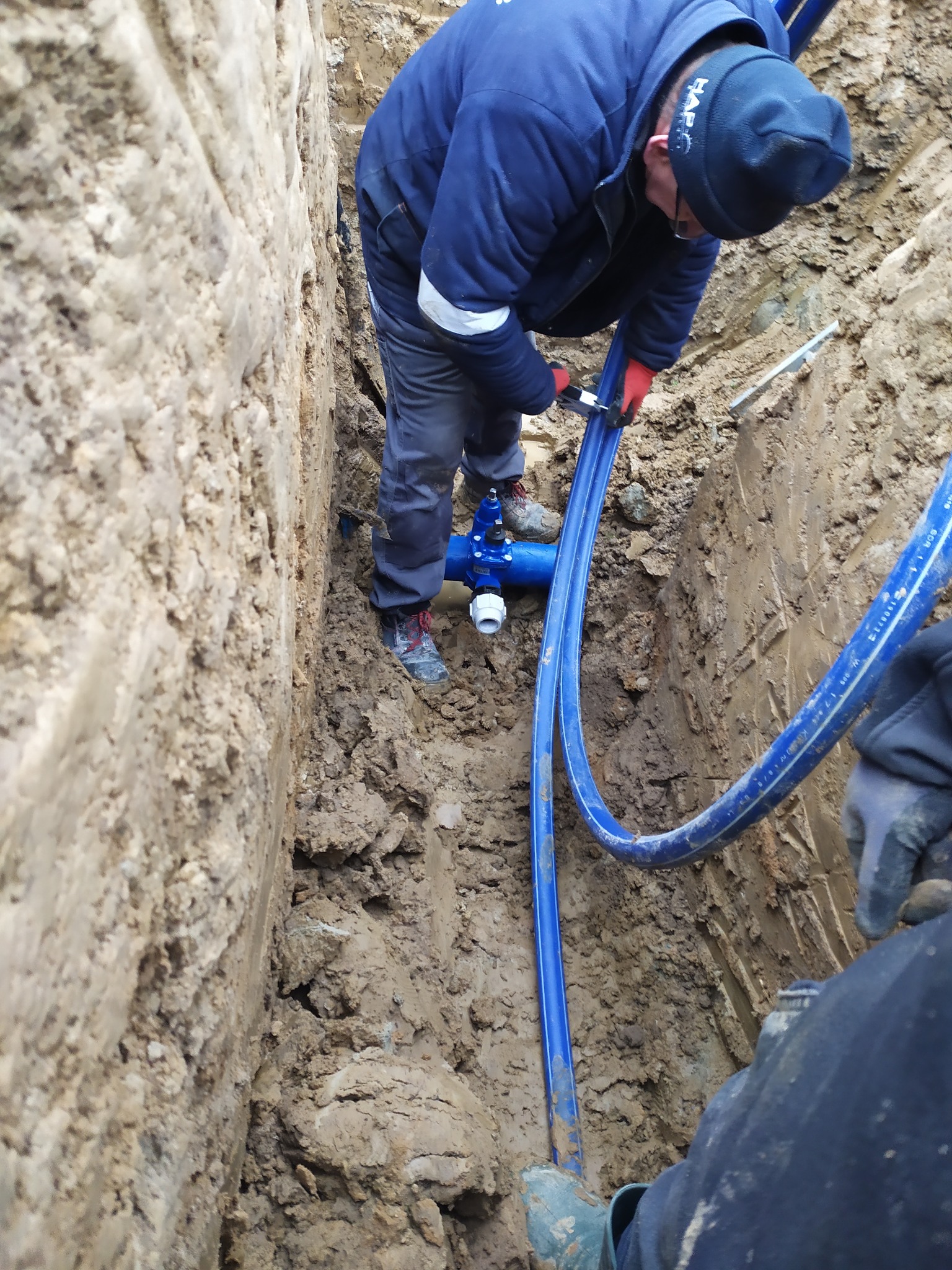 Pracownik w wykopie łączy niebieskie rury z armaturą. Widoczne ściany wykopu z ziemi i gliny, narzędzia oraz fragmenty ubioru innych osób.