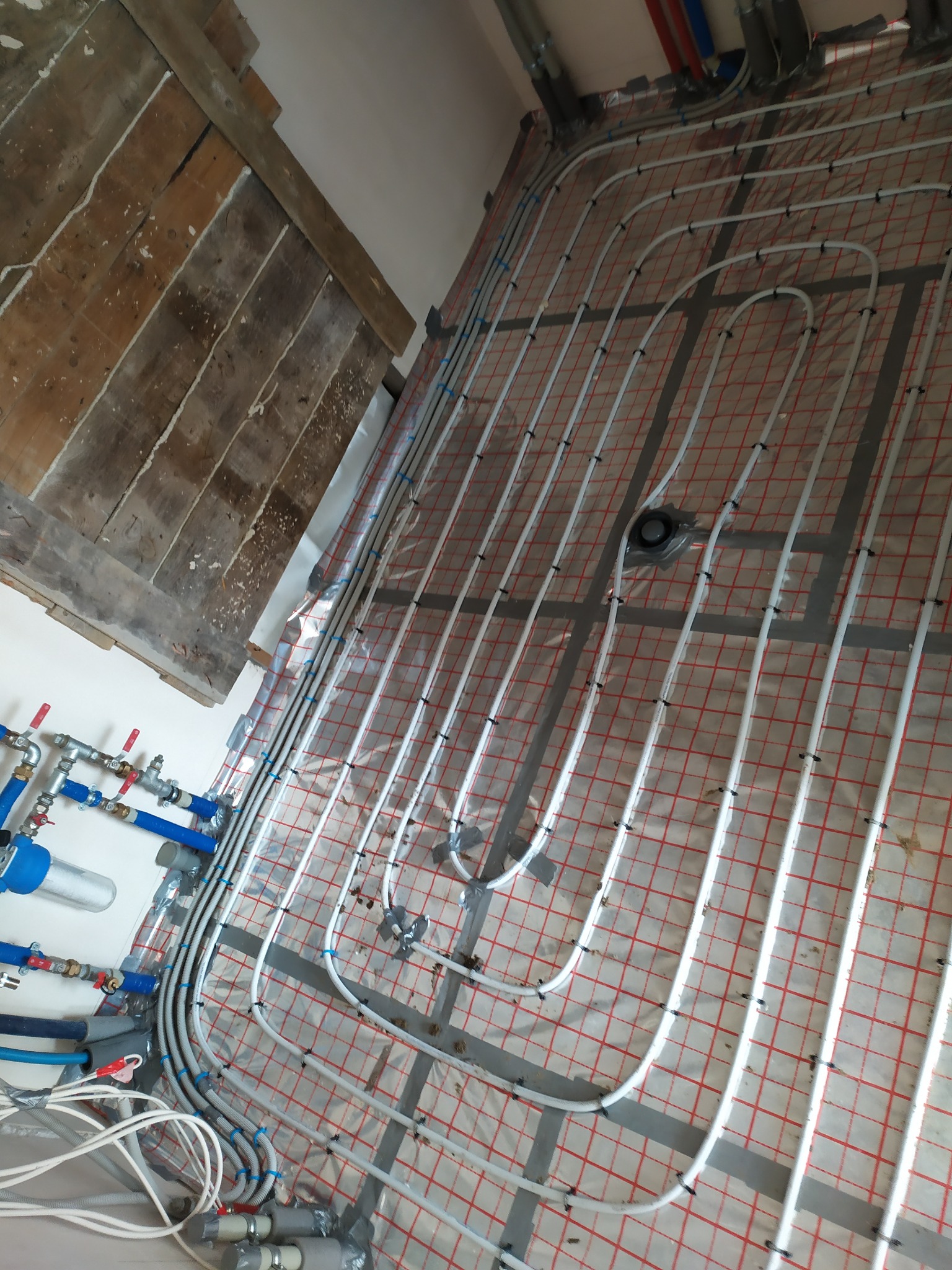 Instalacja ogrzewania podłogowego z białych rur na folii z czerwoną kratką, widoczne elementy hydrauliczne i fragment drewnianej podłogi w tle.