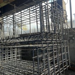 JFK GROUP KOWALCZYK Sp. z o.o. - Klatki z drutu zbrojeniowego ułożone jedna na drugiej, widoczne w hali produkcyjnej z oknami w tle. Elementy konstrukcyjne do fundamentów.