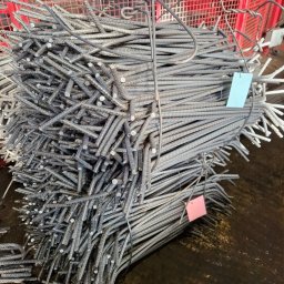 JFK GROUP KOWALCZYK Sp. z o.o. - Wiązka stalowych prętów zbrojeniowych z niebieską i różową etykietą, ułożona na ciemnym podłożu, z fragmentem czerwonej konstrukcji kratowej w tle.