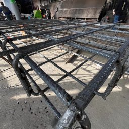 JFK GROUP KOWALCZYK Sp. z o.o. - Gotowa klatka zbrojeniowa z żebrowanych prętów, połączonych drutem wiązałkowym, widoczna z bliska na tle hali produkcyjnej. Element konstrukcyjny do fundamentów.