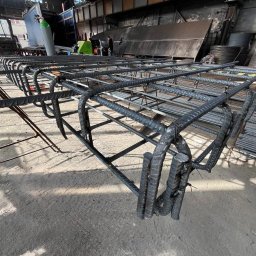 JFK GROUP KOWALCZYK Sp. z o.o. - Szkielet z prętów zbrojeniowych, połączonych drutem, tworzący przestrzenną konstrukcję w hali produkcyjnej. Widoczne elementy maszyny i składowane pręty.