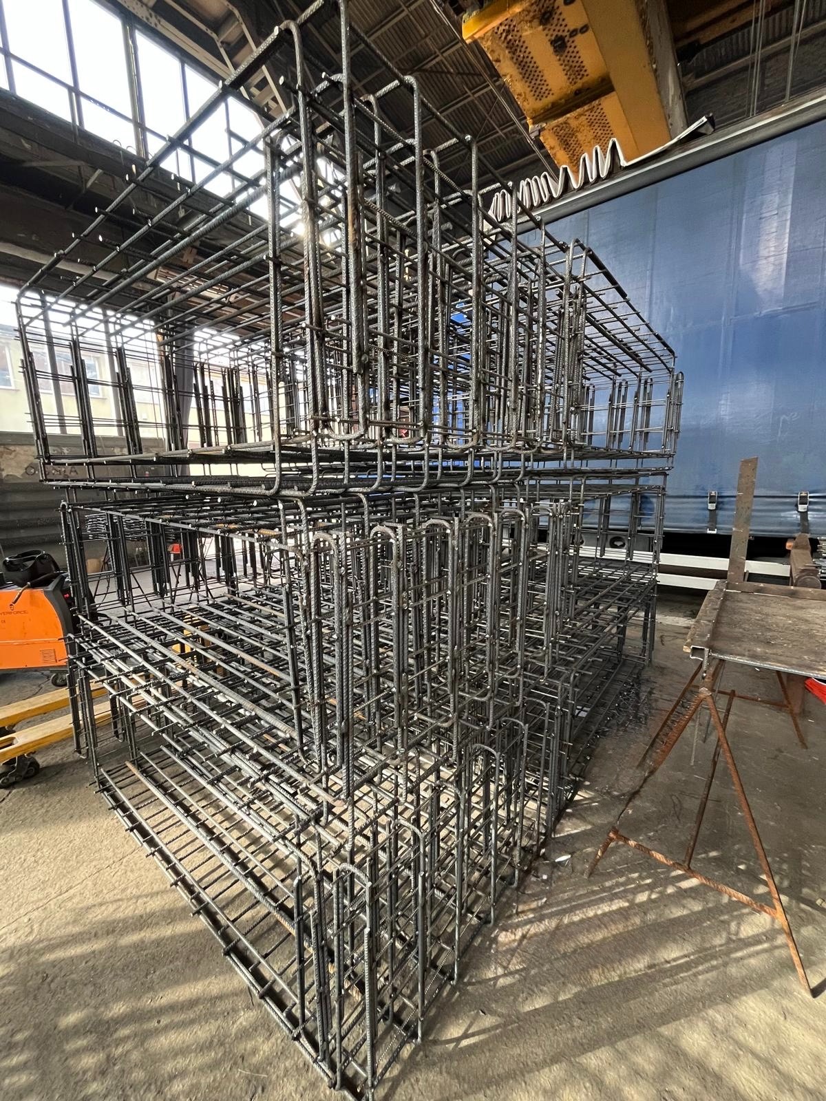 Klatki zbrojeniowe ułożone jedna na drugiej w hali produkcyjnej, widoczne elementy konstrukcyjne budynku i wózek widłowy w tle. Szare, surowe wykończenie.