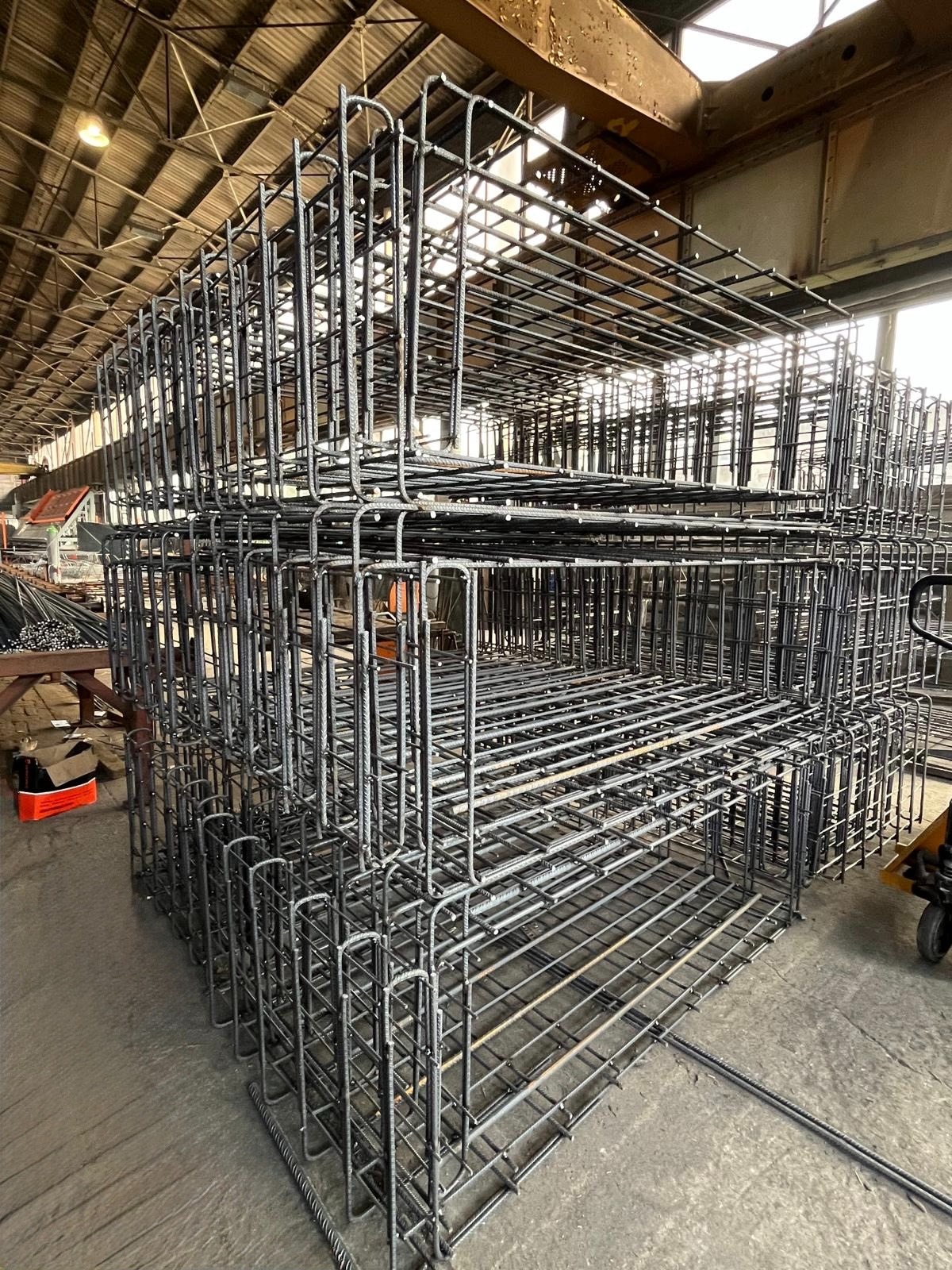 Klatki z prętów zbrojeniowych ułożone w stos na hali produkcyjnej, widoczne elementy konstrukcji dachu i oświetlenie przemysłowe.
