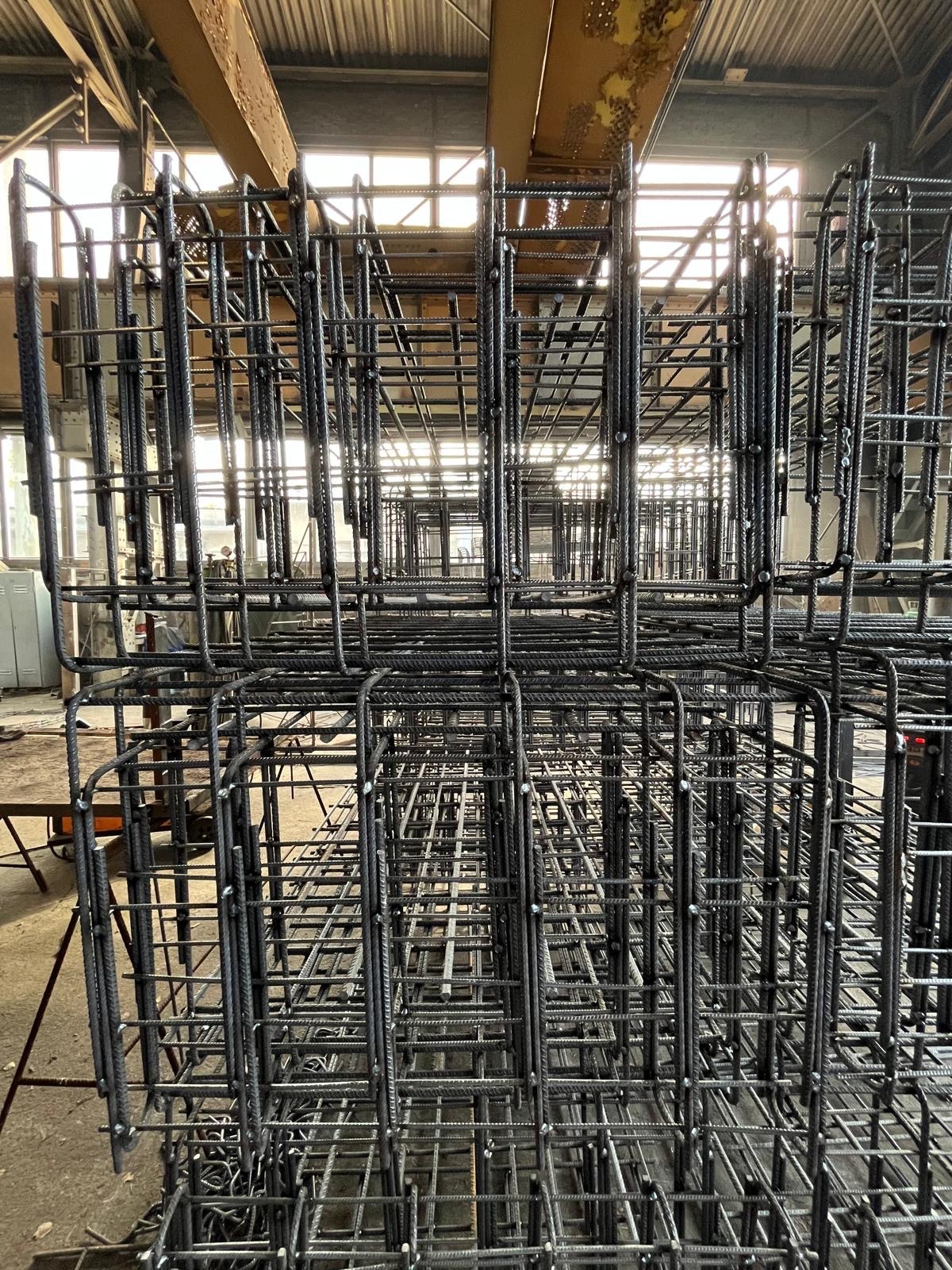 Klatki z prętów żebrowanych, tworzące konstrukcję szkieletową, ustawione w hali produkcyjnej z widocznym oświetleniem górnym i elementami konstrukcyjnymi budynku.