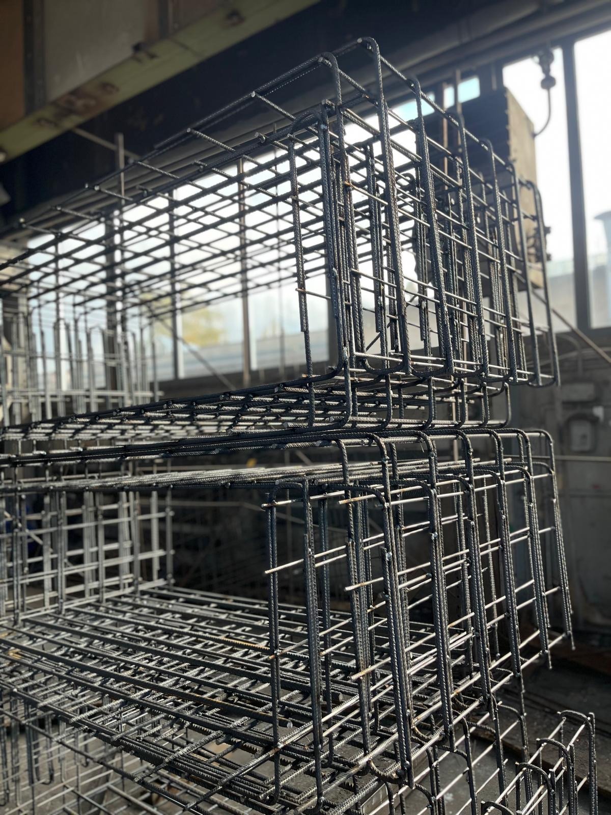 Klatki z drutu zbrojeniowego ułożone jedna na drugiej, widoczne w hali produkcyjnej z oknami w tle. Elementy konstrukcyjne do fundamentów.