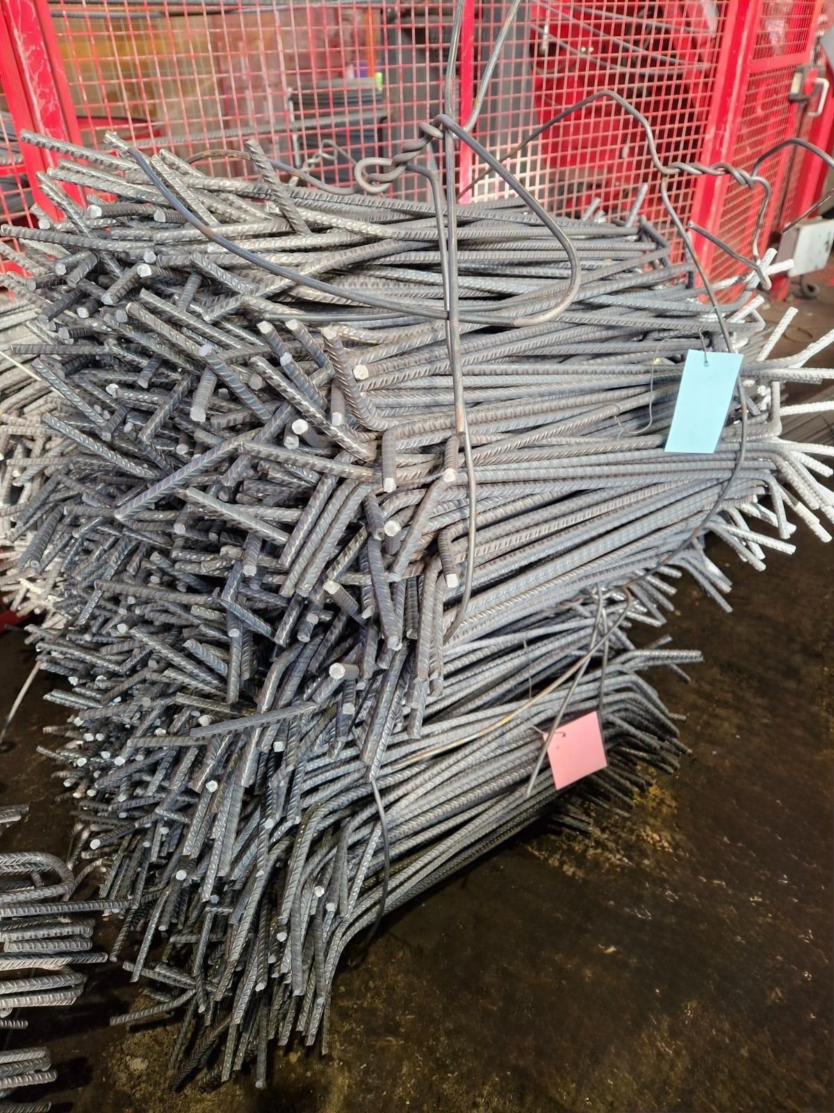 Wiązka stalowych prętów zbrojeniowych z niebieską i różową etykietą, ułożona na ciemnym podłożu, z fragmentem czerwonej konstrukcji kratowej w tle.