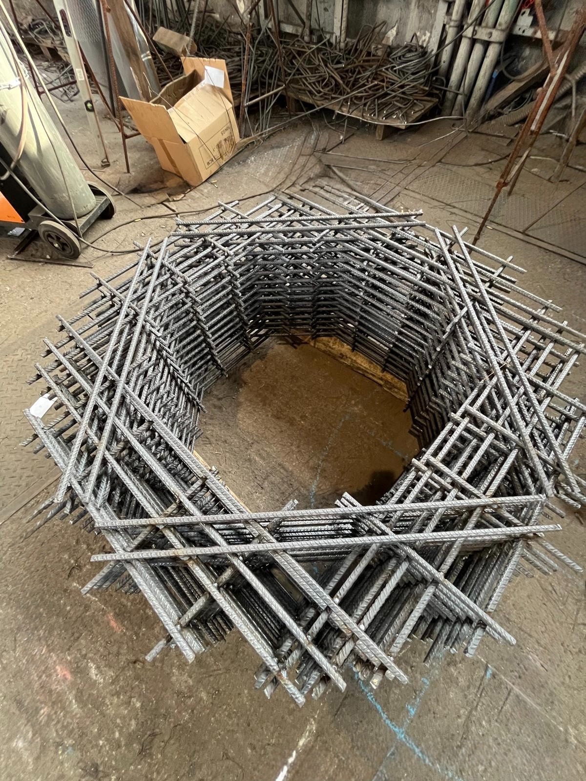 Klatka z prętów żebrowanych, ułożona warstwami w kształt wielokąta z pustym środkiem, na podłodze warsztatu. Widoczne ślady spawania i obróbki.