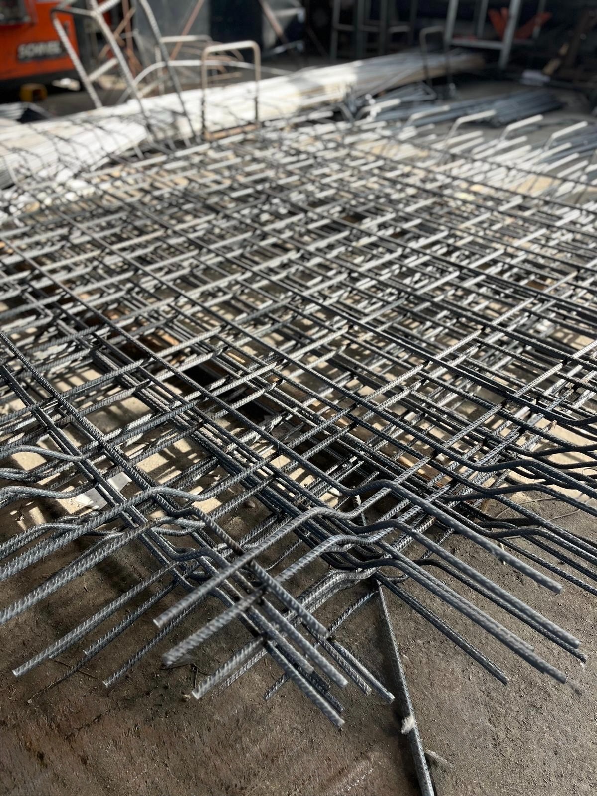 Siatka z prętów żebrowanych ułożona na podłożu, widoczne zagięte końcówki prętów. W tle elementy konstrukcyjne i maszyna budowlana.