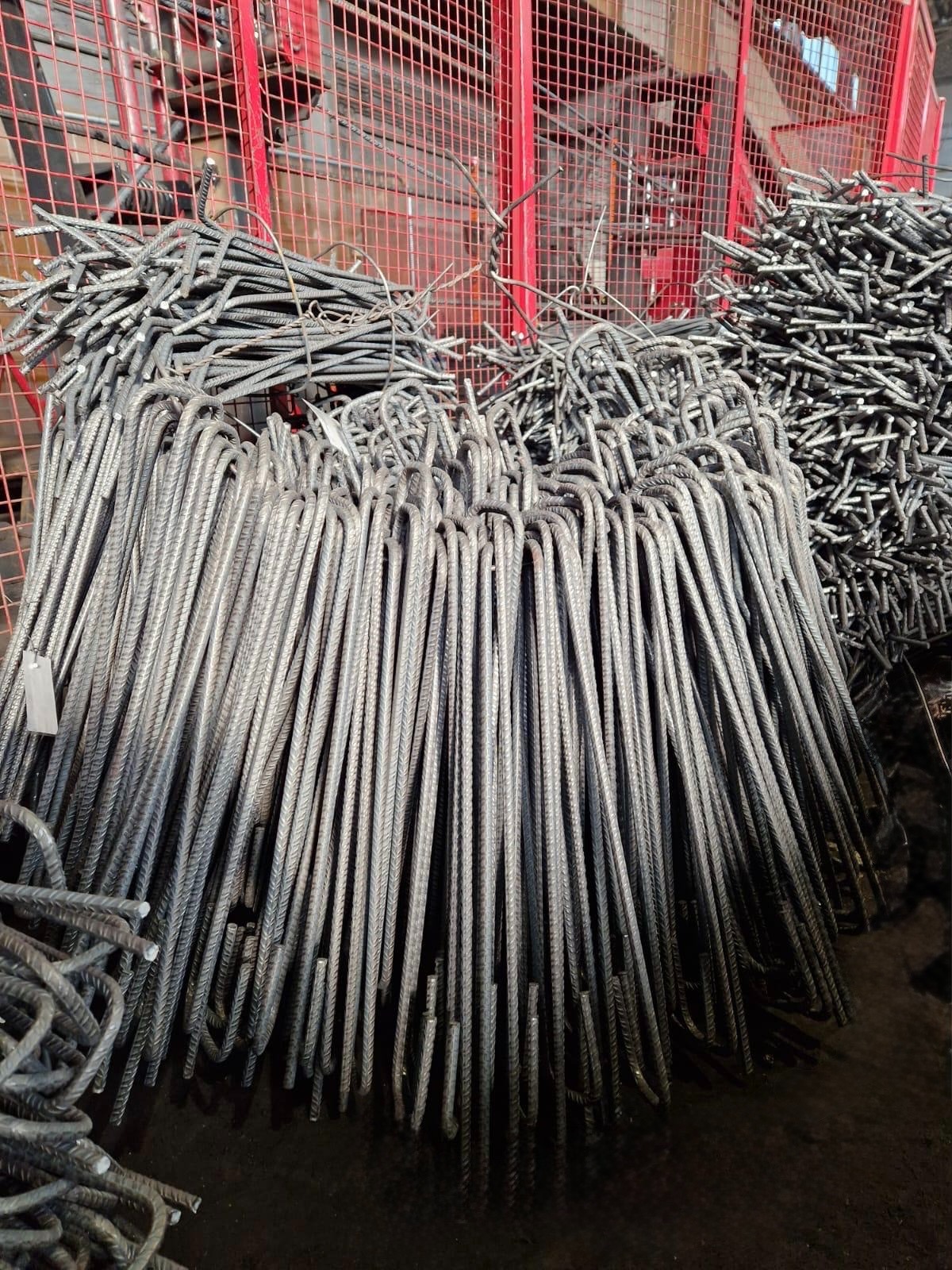 U-kształtne pręty zbrojeniowe ułożone w stos, widoczne żebrowania, w tle czerwona krata i inne pręty. Magazyn materiałów budowlanych.