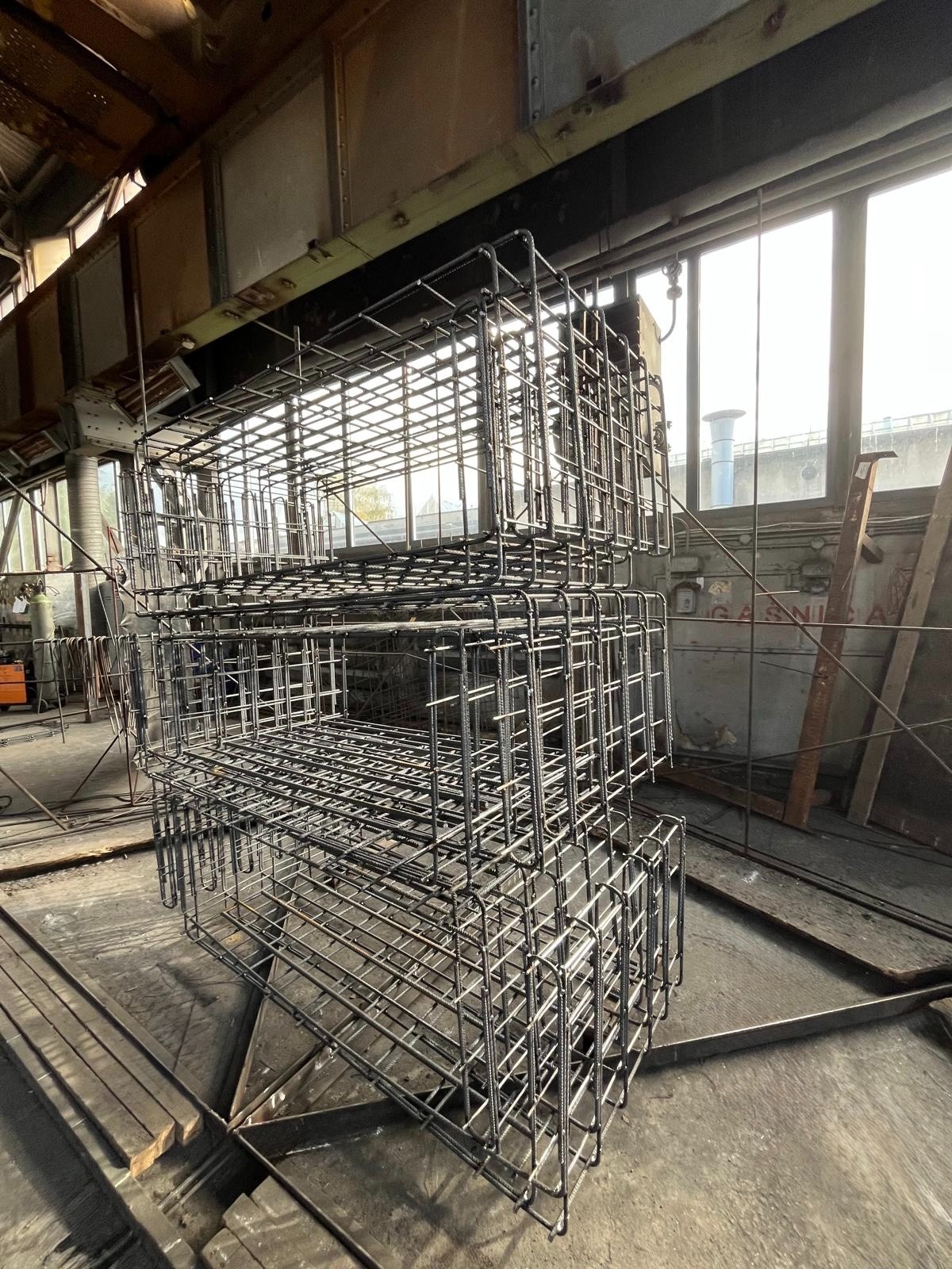 Klatki z prętów zbrojeniowych ułożone jedna na drugiej w hali produkcyjnej. Widoczne okna w tle i elementy konstrukcji hali. Surowy, industrialny charakter.