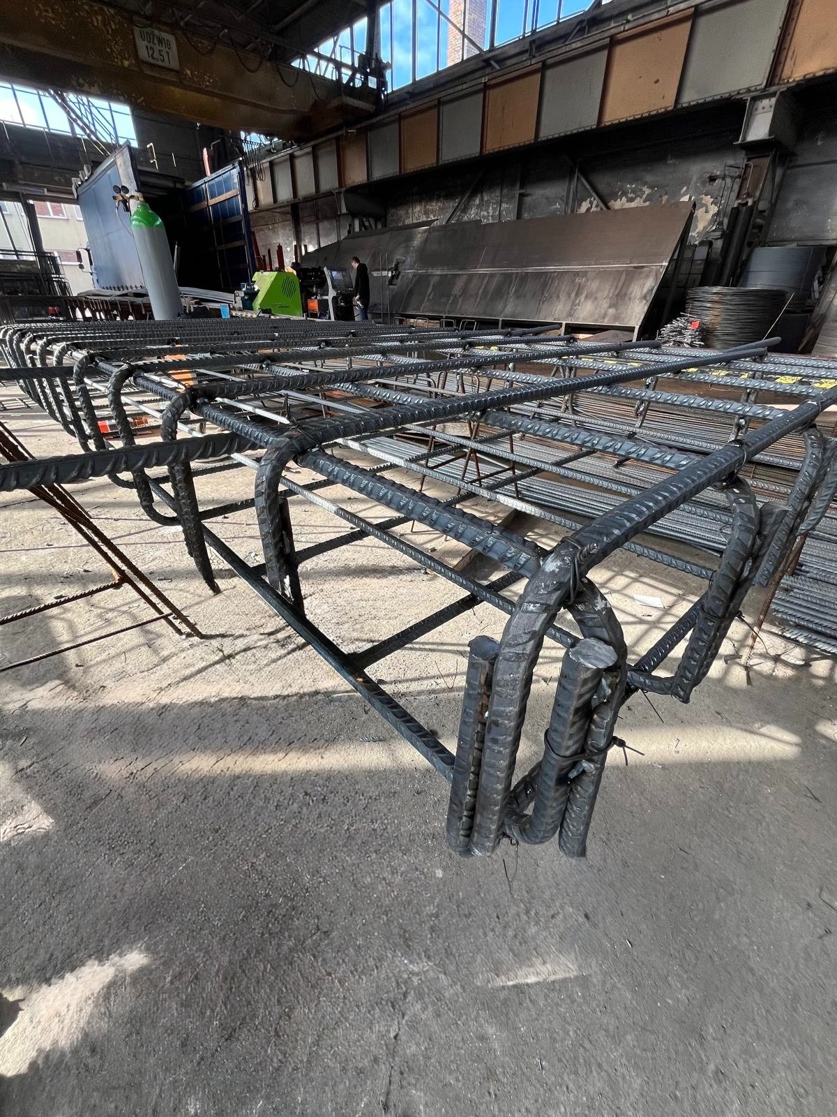 Szkielet z prętów zbrojeniowych, połączonych drutem, tworzący przestrzenną konstrukcję w hali produkcyjnej. Widoczne elementy maszyny i składowane pręty.