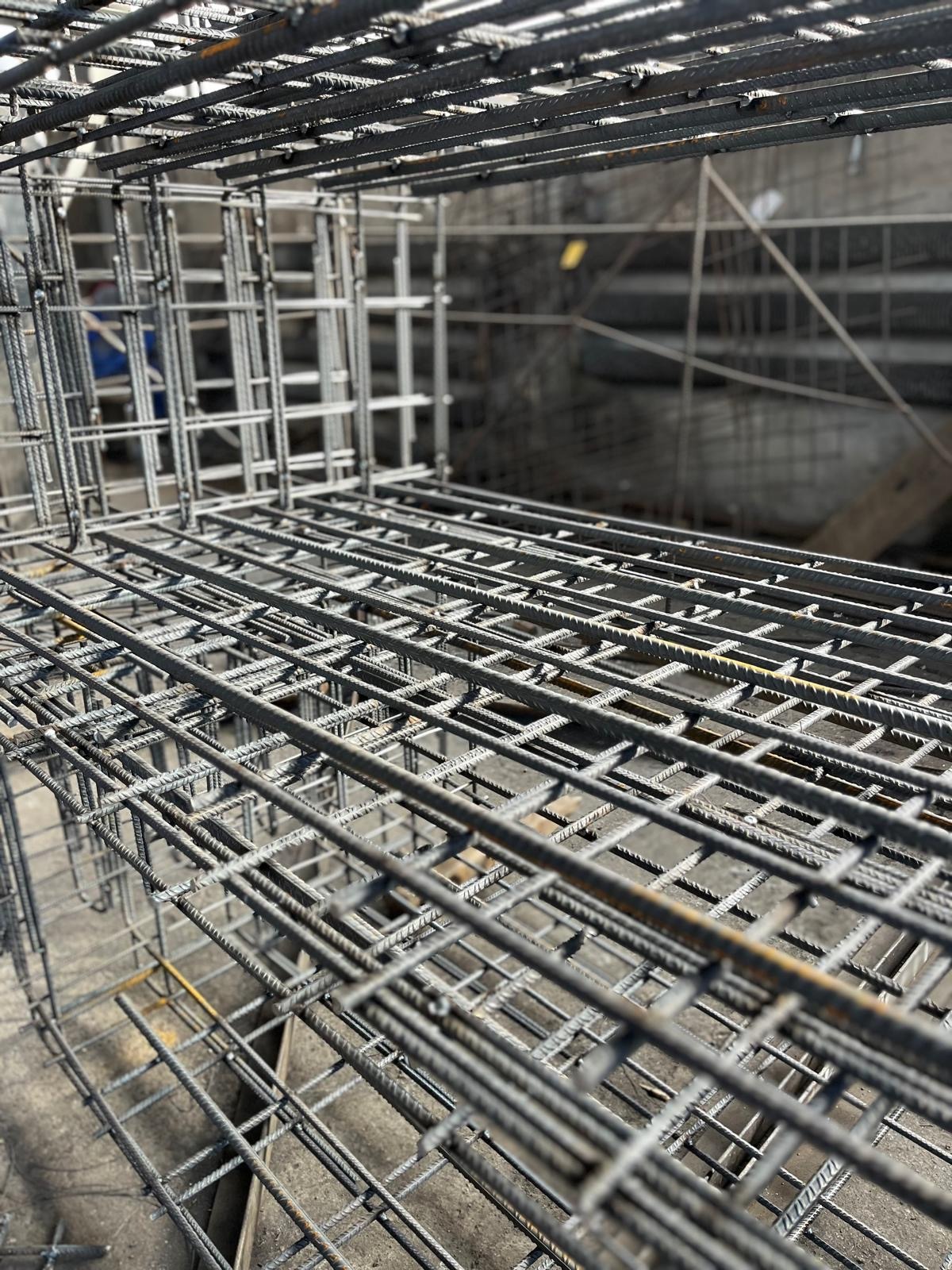 Siatka zbrojeniowa ułożona w przestrzenną konstrukcję, widoczna faktura stali i zgrzewy, w tle elementy konstrukcyjne budynku w surowym stanie.