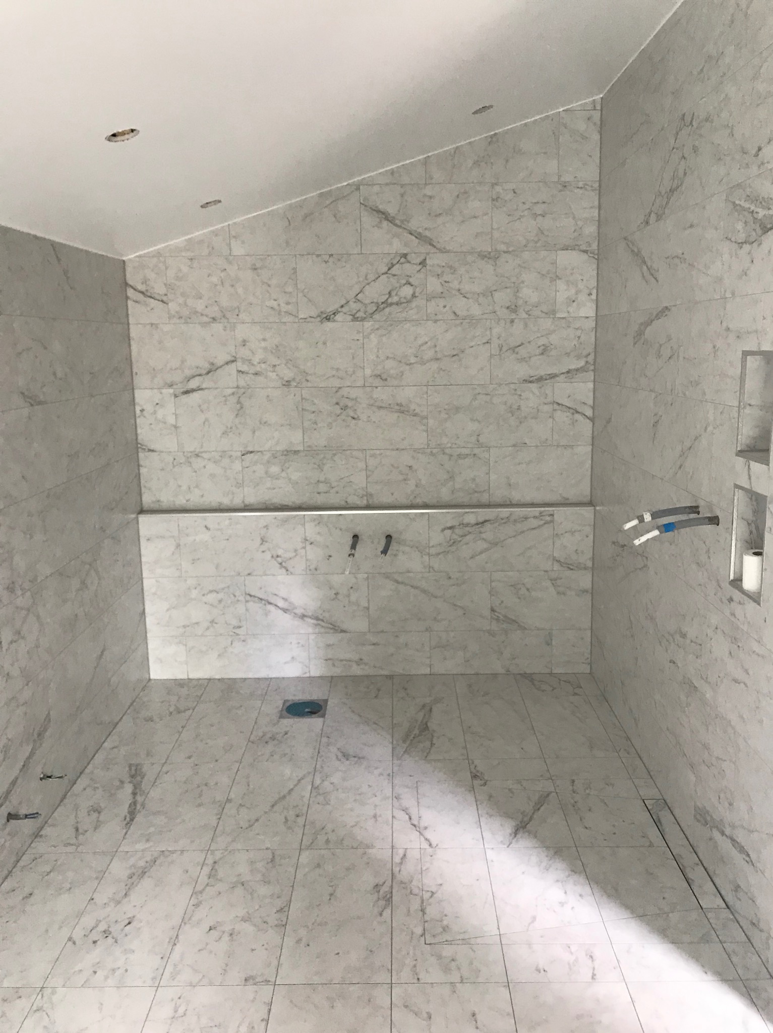 Wykończona łazienka z marmurowymi płytkami, widoczne instalacje wodne i odpływ liniowy. Półki w ścianie z rolką papieru toaletowego. Otwory w suficie pod oświetlenie.