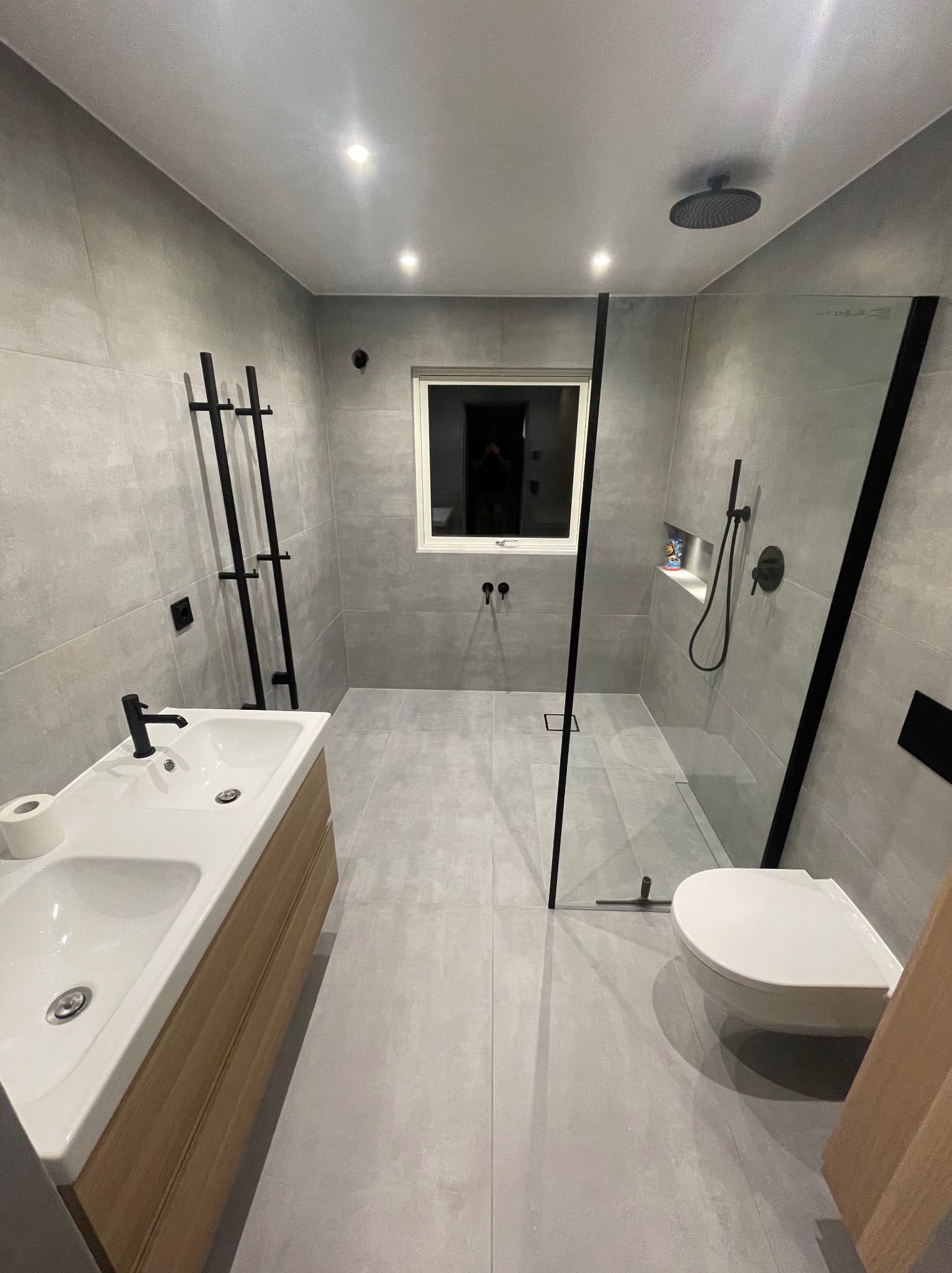 Nowoczesna łazienka w szarościach z kabiną typu walk-in, umywalką nablatową z drewnianą szafką i czarnymi akcentami, oświetlona punktowo.