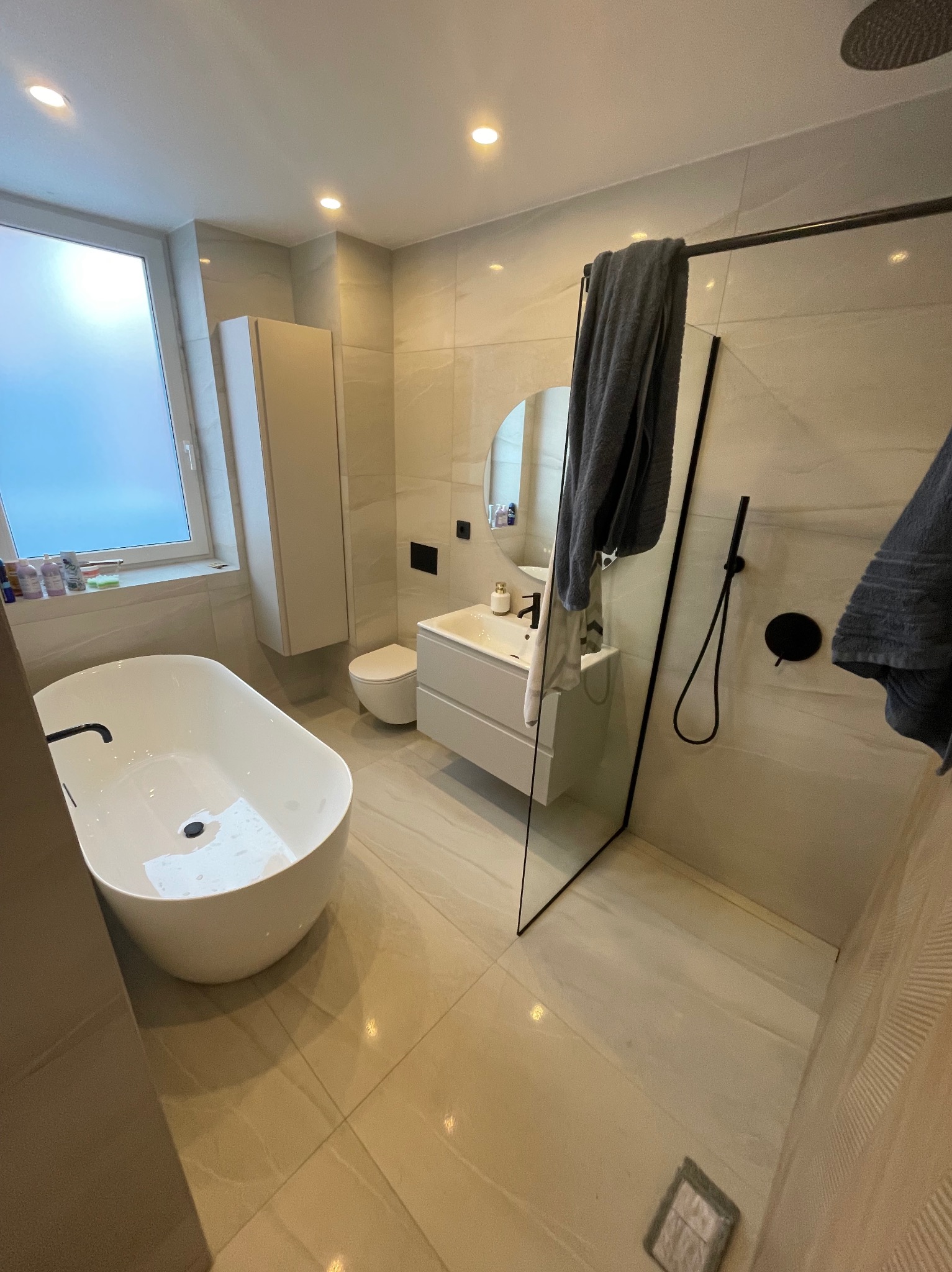 Jasna łazienka z wolnostojącą wanną, kabiną prysznicową ze szklaną ścianką i czarną armaturą. Na ścianach płytki imitujące marmur. Minimalistyczny styl.