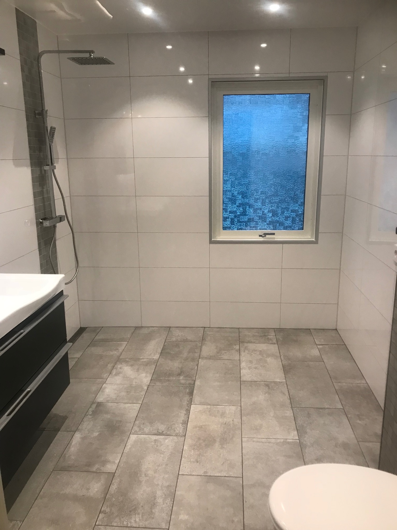 Jasna łazienka z białymi płytkami ściennymi, szarą podłogą z płytek imitujących kamień i oknem z teksturowaną szybą. Widoczna kabina prysznicowa i umywalka z szafką.