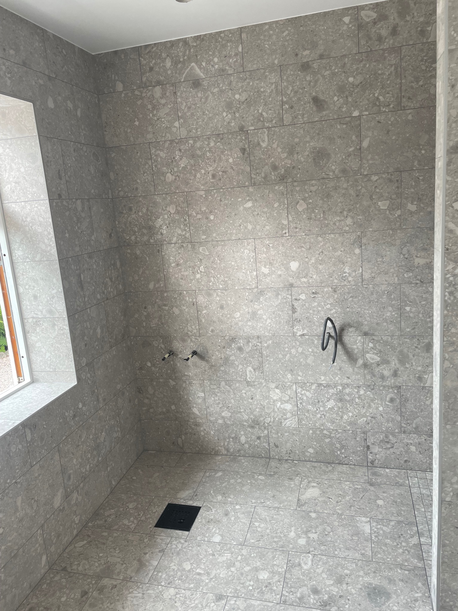 Wykończona kabina prysznicowa z szarymi płytkami imitującymi kamień, widoczne przyłącza wodne i odpływ liniowy. Minimalistyczny design z oknem po lewej stronie.